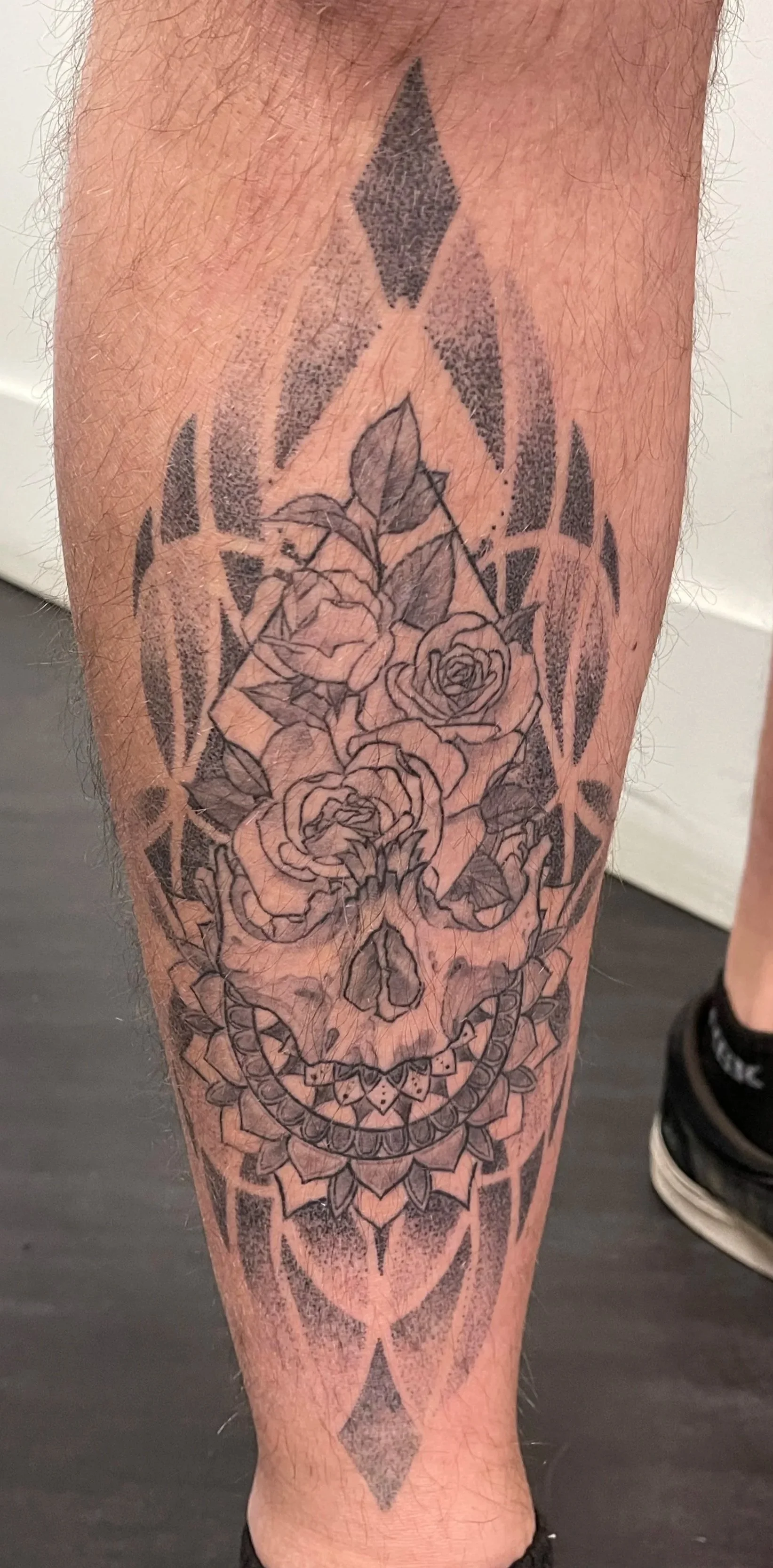 Tattoo of a skull with roses on the forehead, surrounded by geometric patterns on the lower leg.