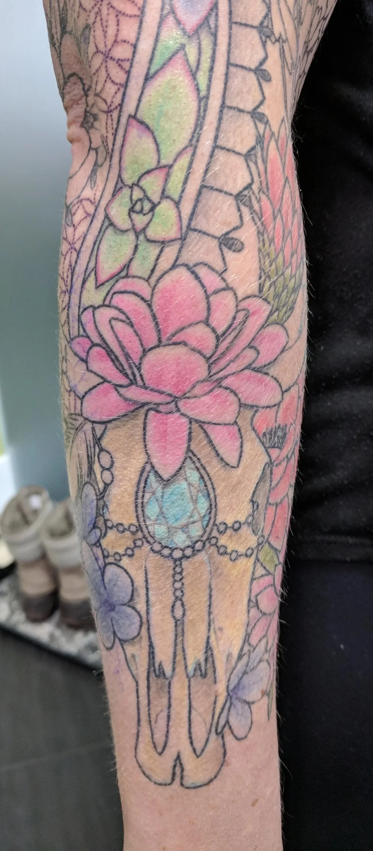 Colorful tattoo on arm featuring a pink flower, a hanging pendant with a blue gemstone, and other floral and decorative elements.