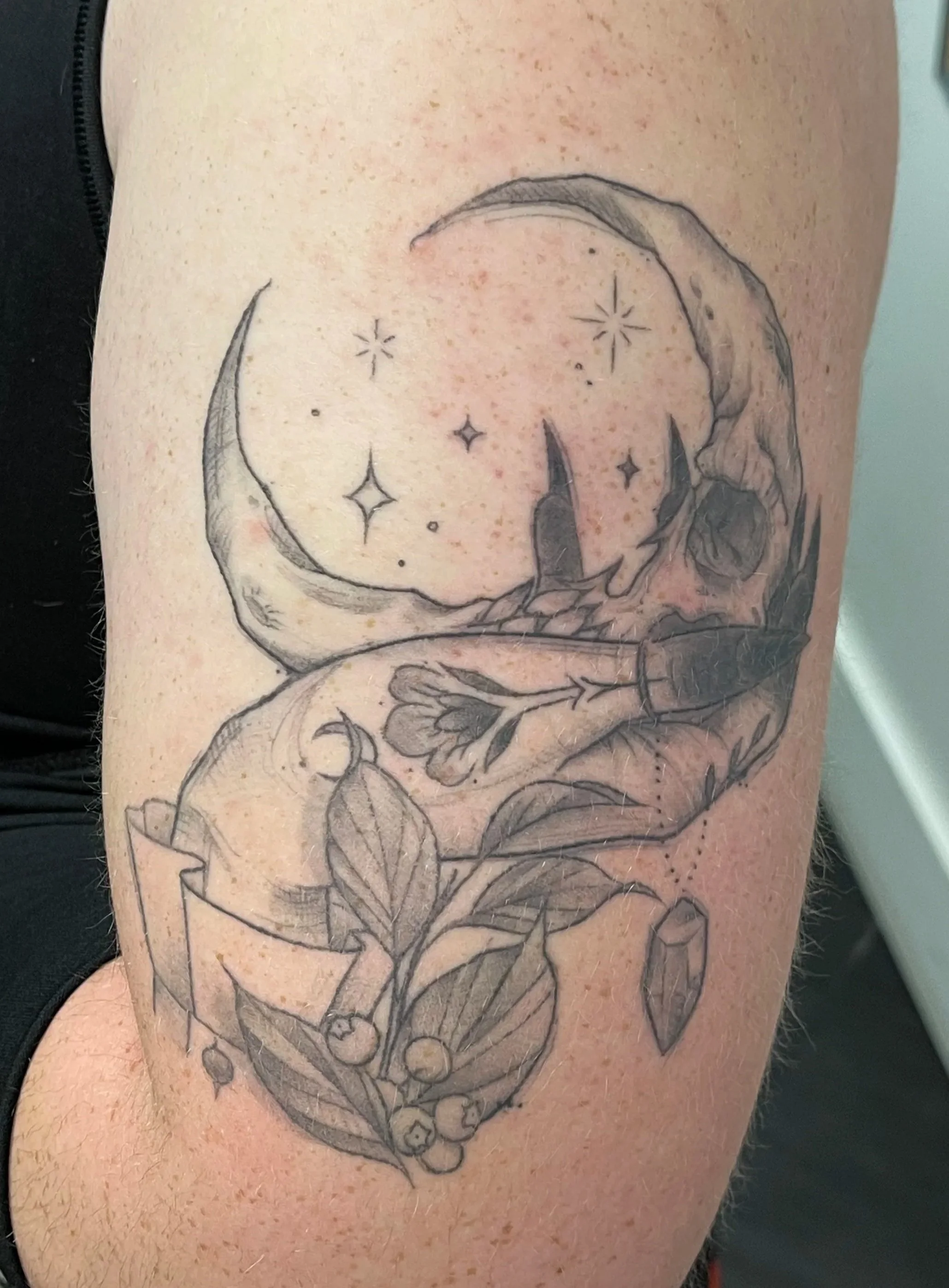 Black and gray tattoo on upper arm featuring a crescent moon, a skull, a butterfly, leaves, berries, stars, and a crystal.