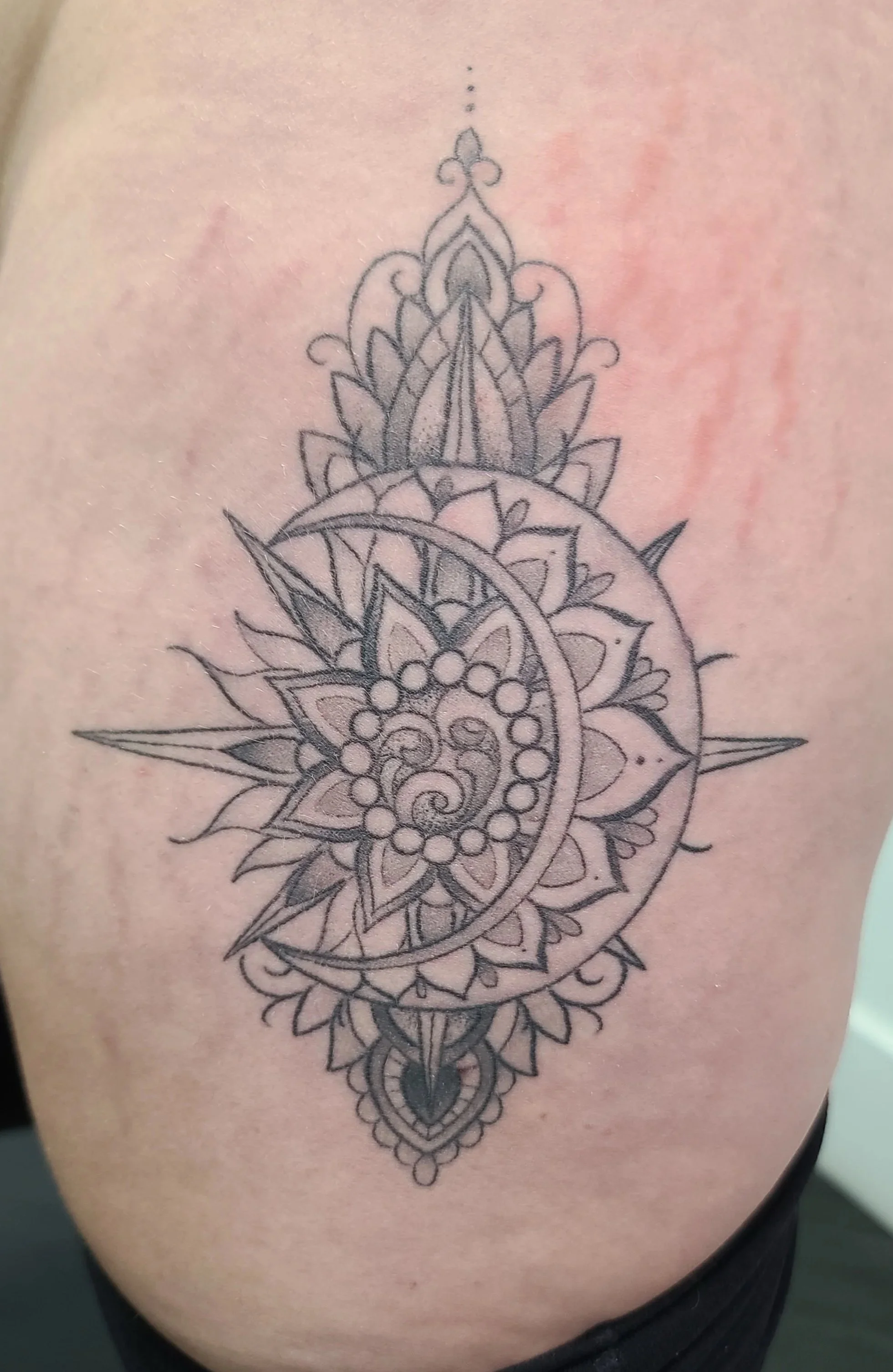 Black outline tattoo of intricate mandala design with a crescent moon and spiral pattern in the center, surrounded by symmetrical floral and geometric patterns.