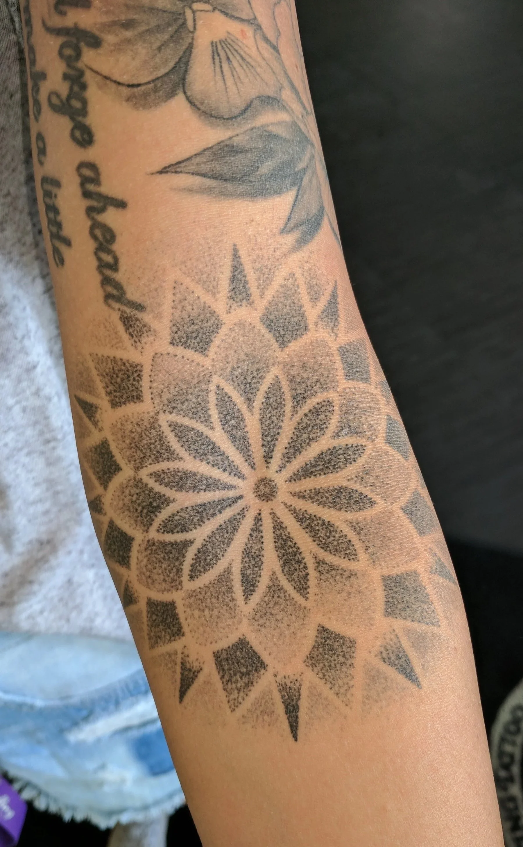 A tattoo on a person's forearm featuring a large geometric floral design in black ink, with part of a floral tattoo and cursive text above it.