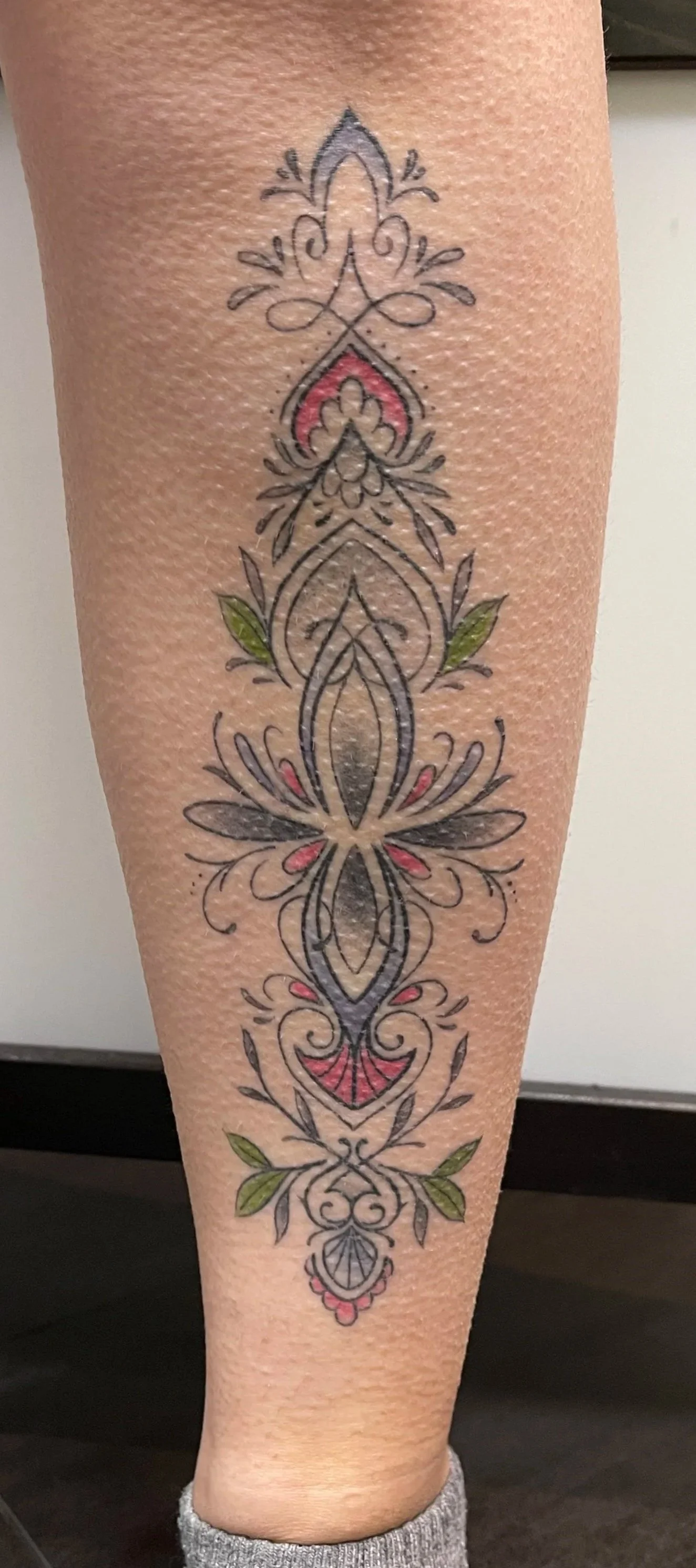Colorful tattoo with intricate black outlines, green leaves, and pink accents on a person's lower leg.