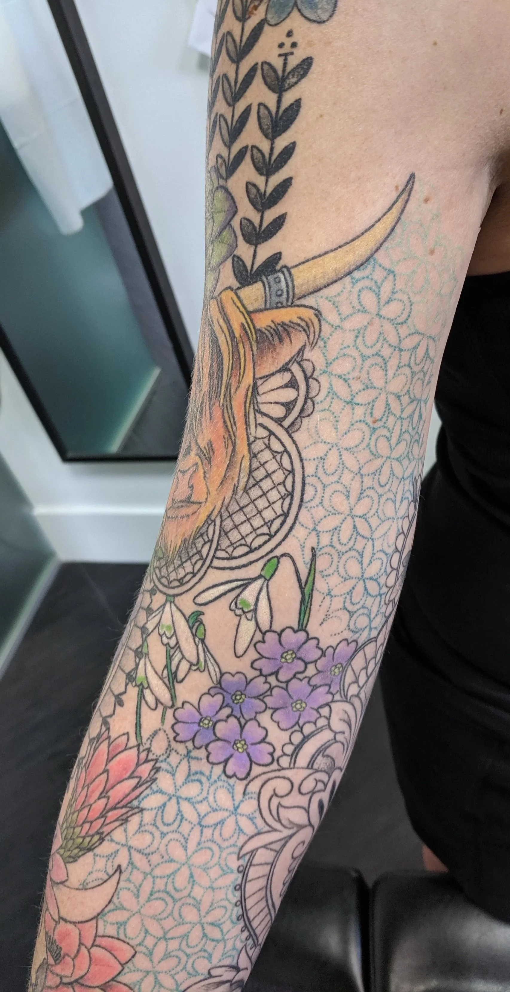 Close-up of a person's arm with a detailed colorful tattoo featuring flowers, a dreamcatcher, and a serpent.