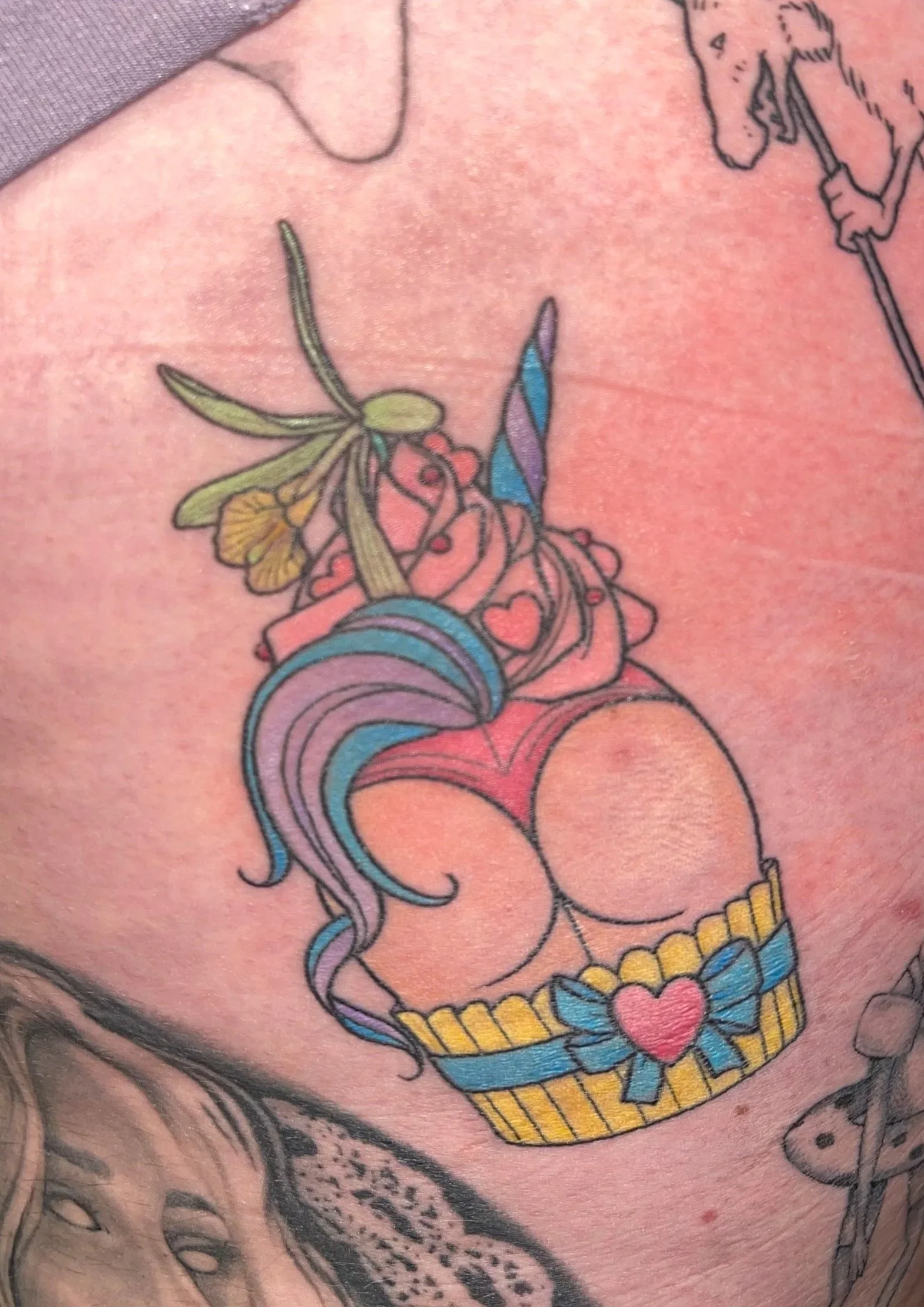 Tattoo of a cupcake with colorful icing, hearts, a butterfly, and a yellow ribbon. Other tattoos visible include a unicorn, a woman’s face, a frog, and a bird.