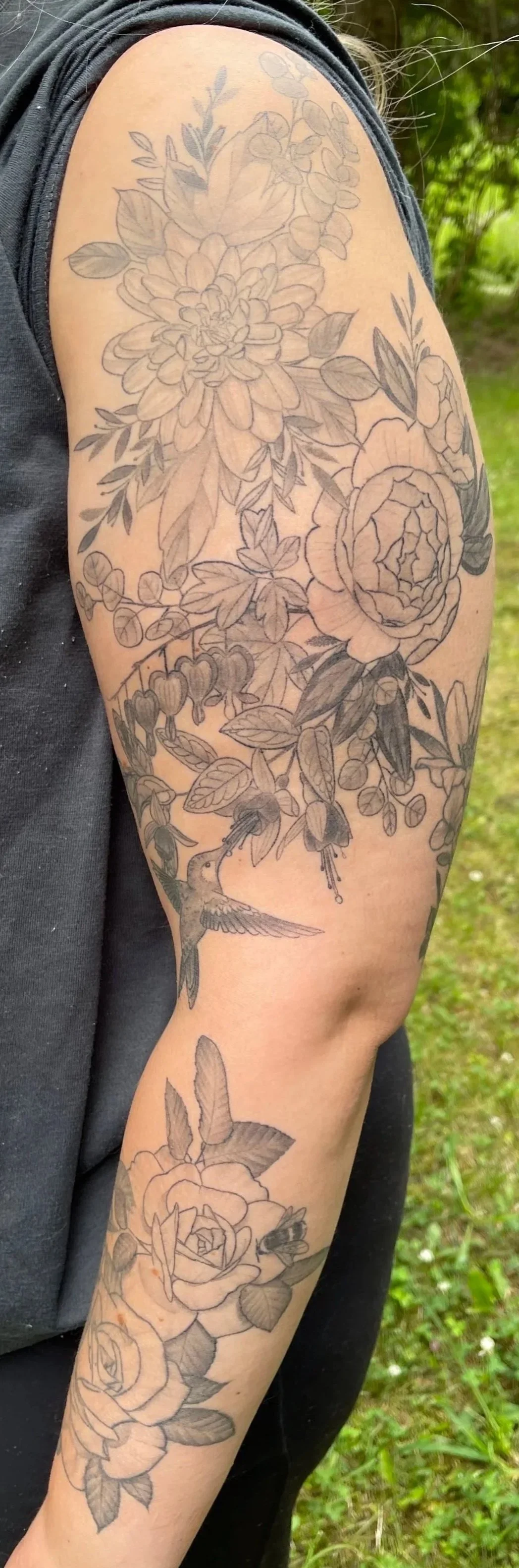 Close-up of a woman's arm with detailed black and gray floral tattoos, including roses, leaves, and a hummingbird, outdoors on green grass.