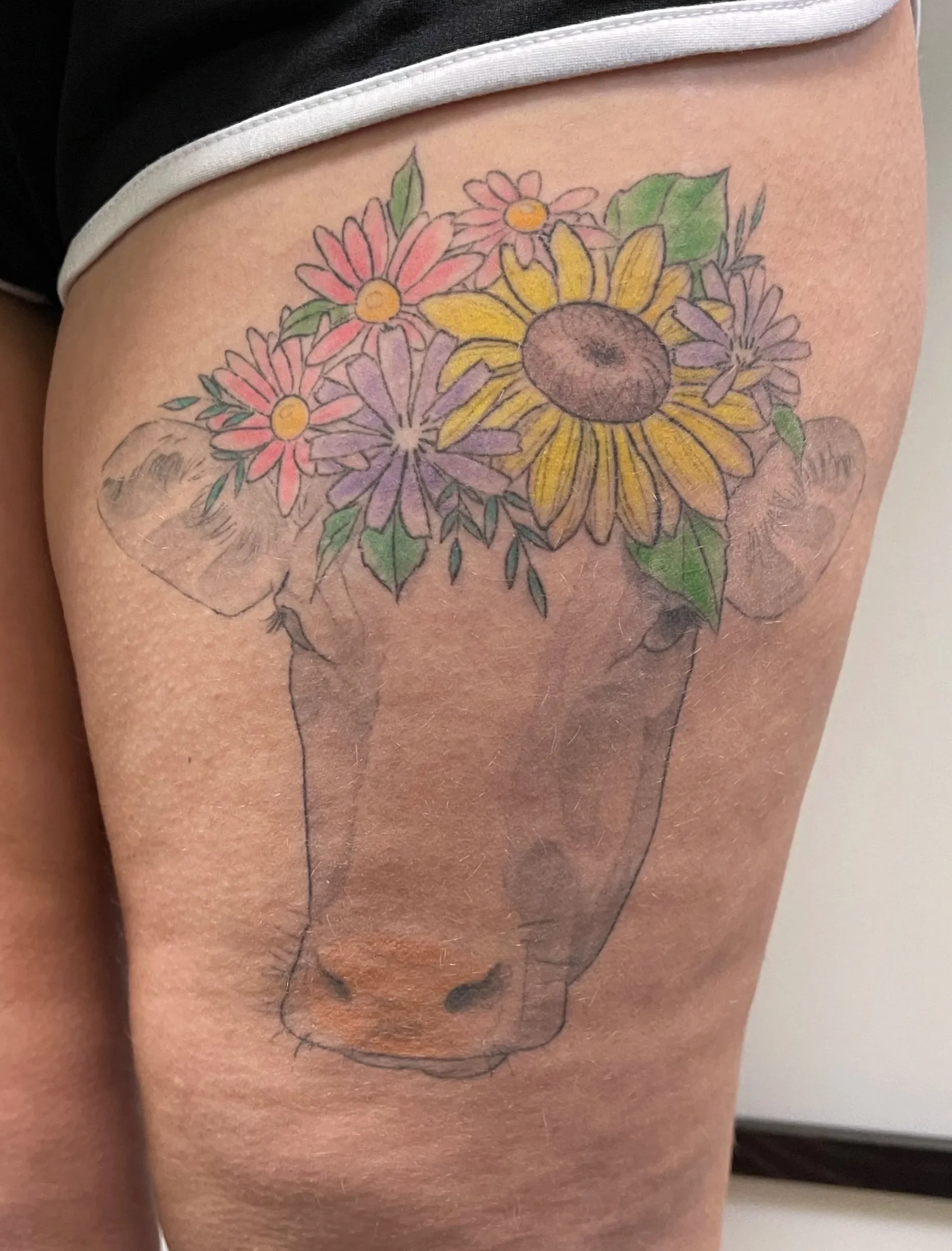 Tattoo of a cow's face with a flower arrangement on its head, on a person's thigh.