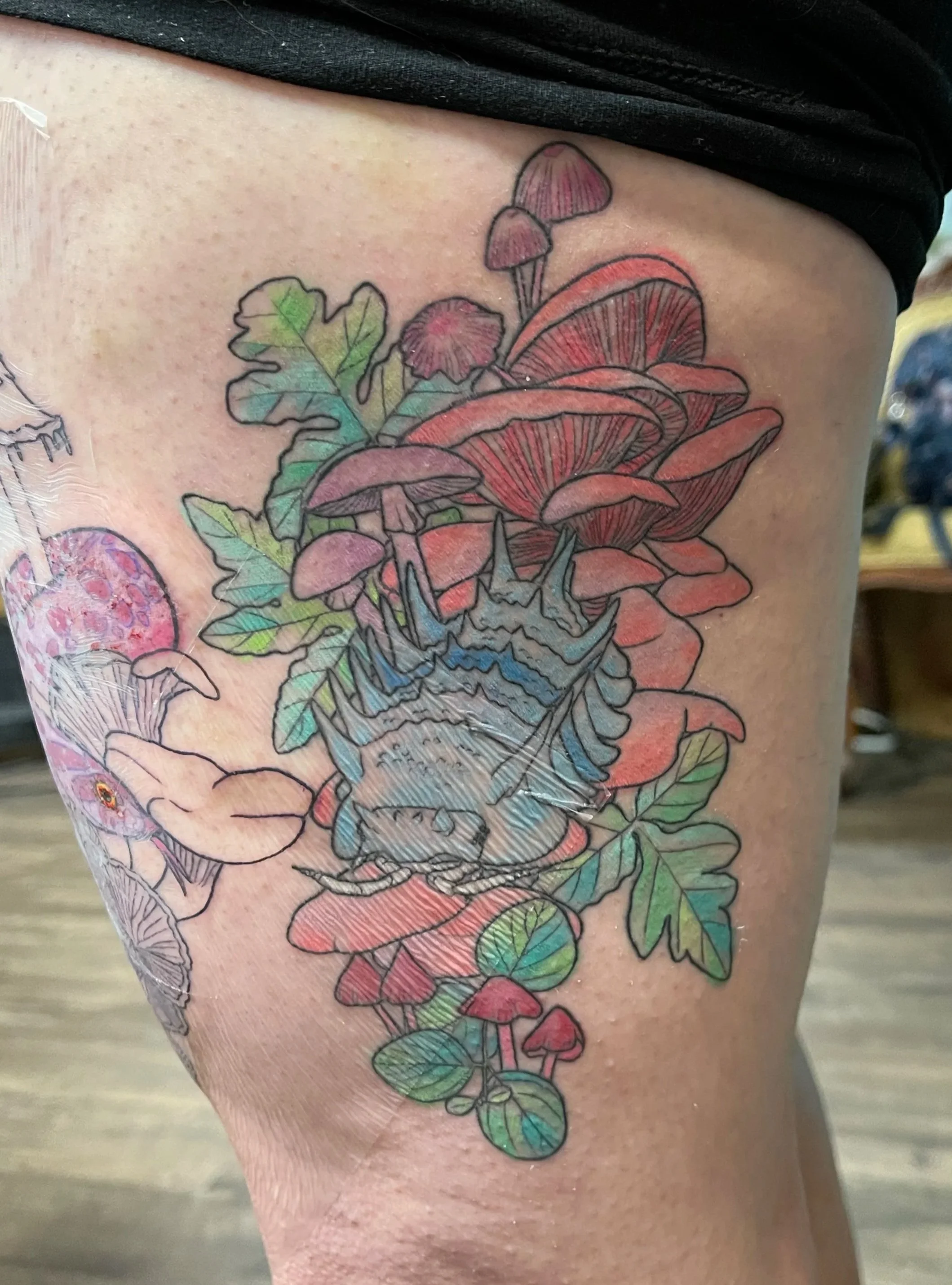 Colorful tattoo of mushrooms, leaves, and a snail on person's thigh.
