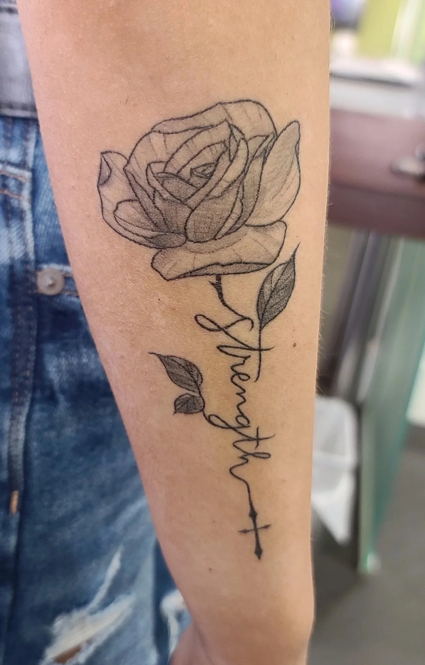 Tattoo of a rose flower with the word 'Afterward' written in cursive and a small cross symbol beneath the script on the person's forearm.