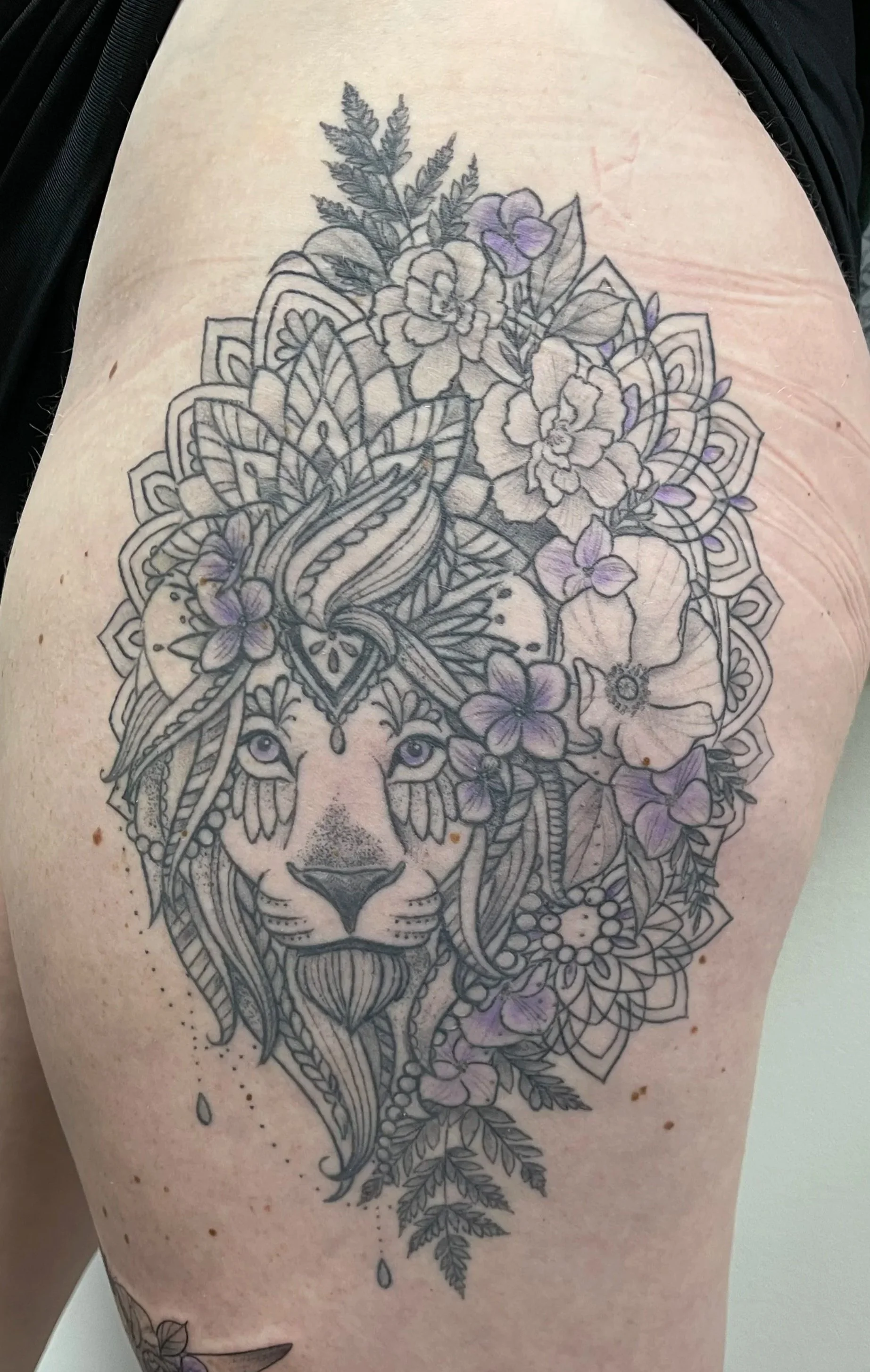 Tattoo of a lion's face surrounded by leaves and flowers, with some parts shaded in purple.