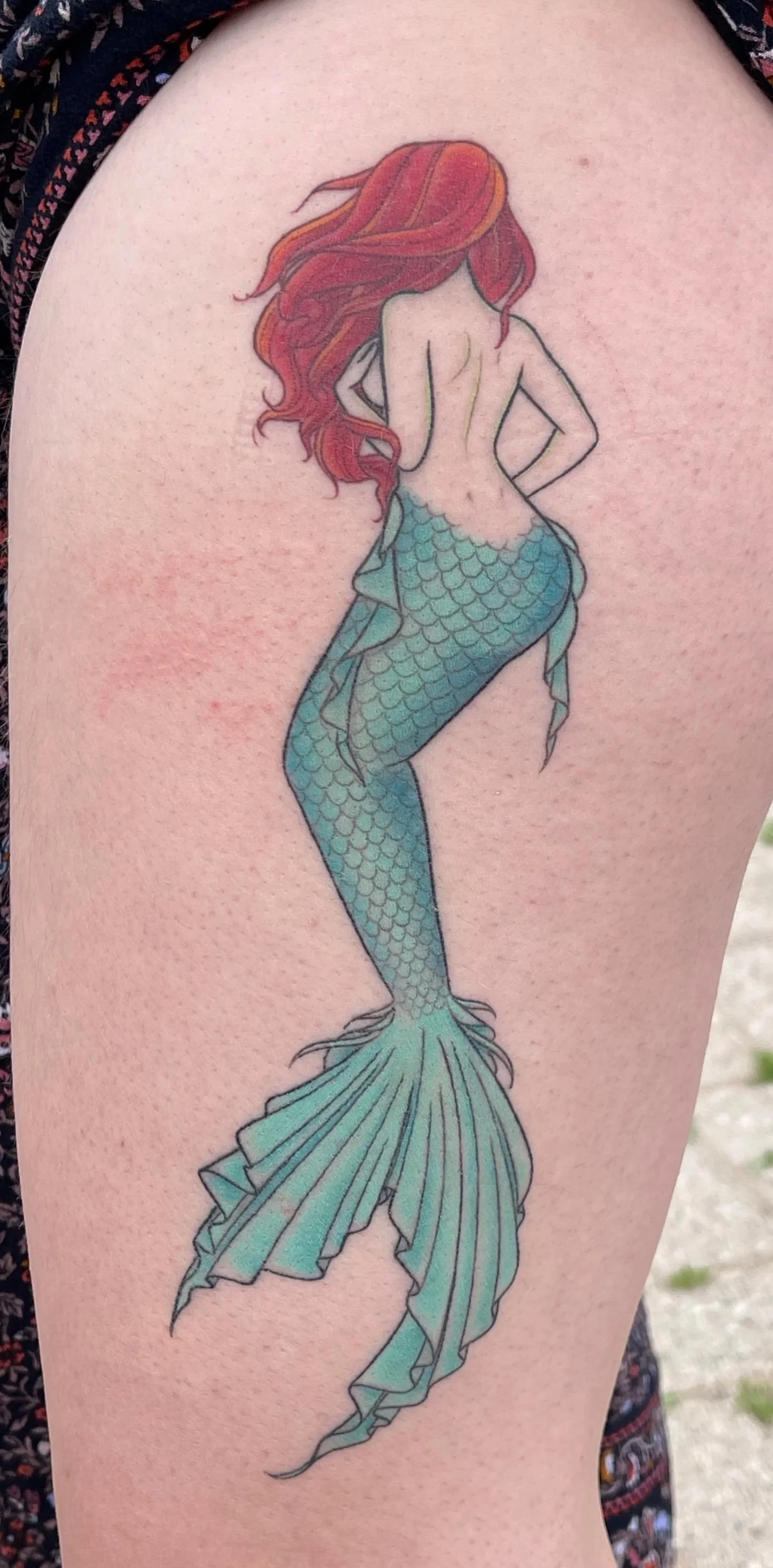 Tattoo of a mermaid with long red hair, facing away, with her hands on her hips, showing her back and a scaled green tail.