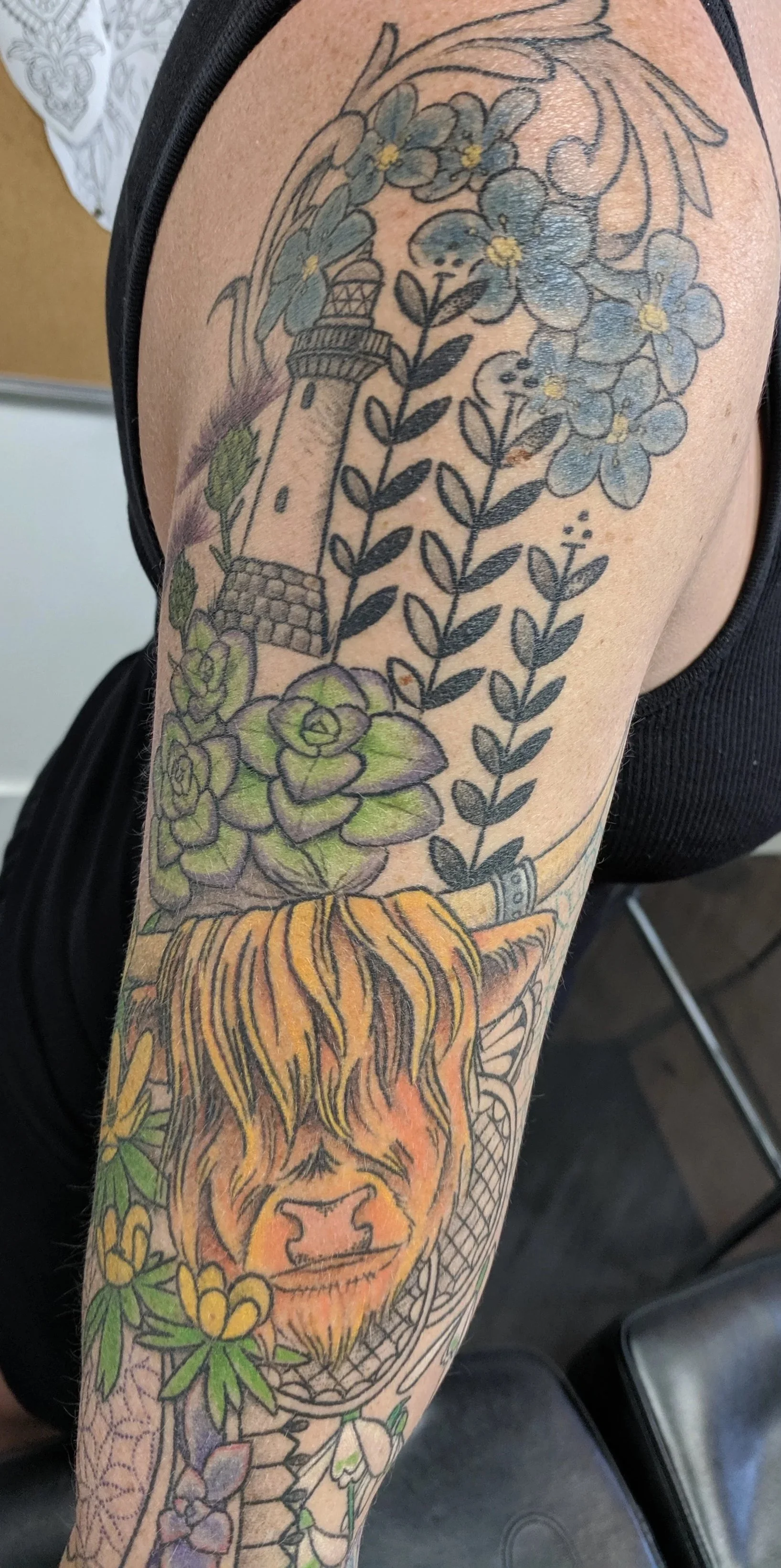 Tattoo of a lighthouse, blue and green flowers, a lion's face, and various plants on a person's upper arm.