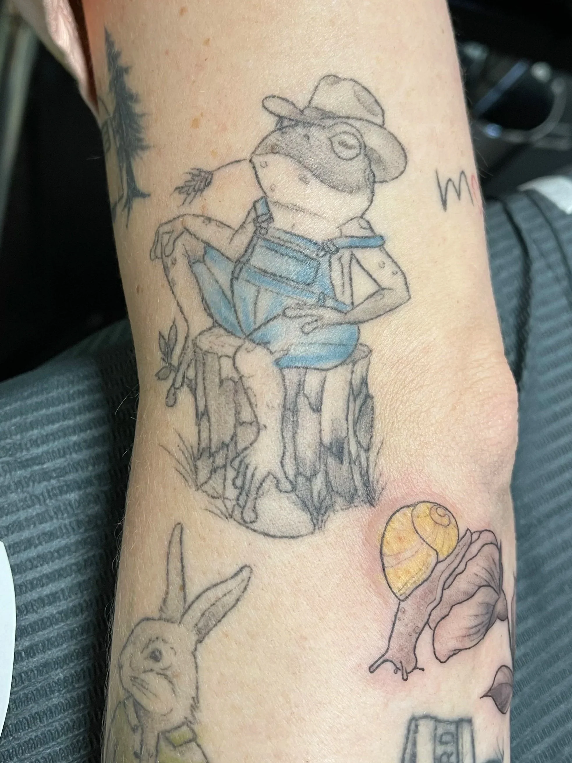 Tattoo of a cartoon mouse with a hat, sitting on a tree stump, holding a leaf in its mouth, dressed in blue overalls.