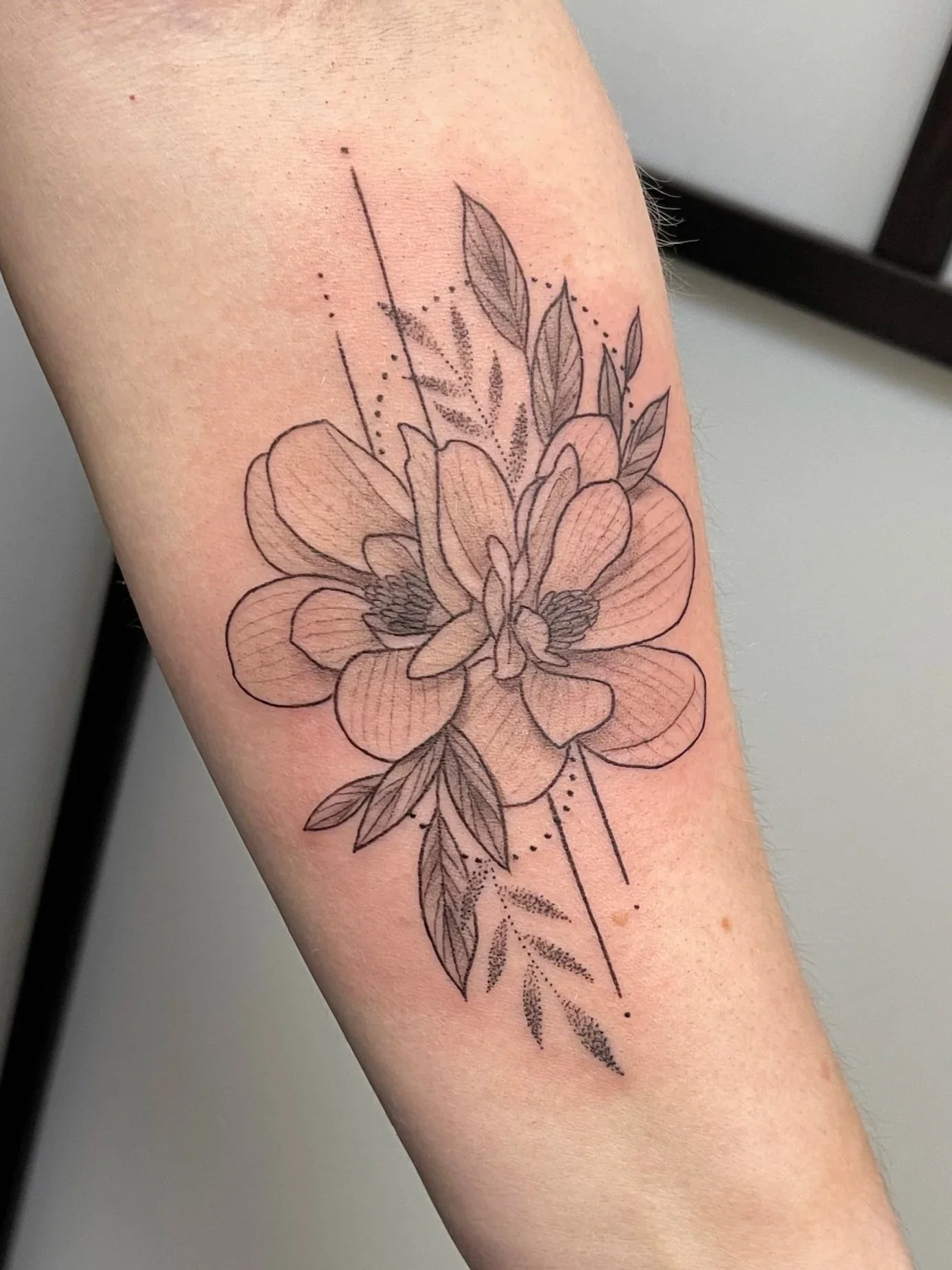 Black and gray floral tattoo on person's forearm featuring a large flower, leaves, and geometric lines and dots.