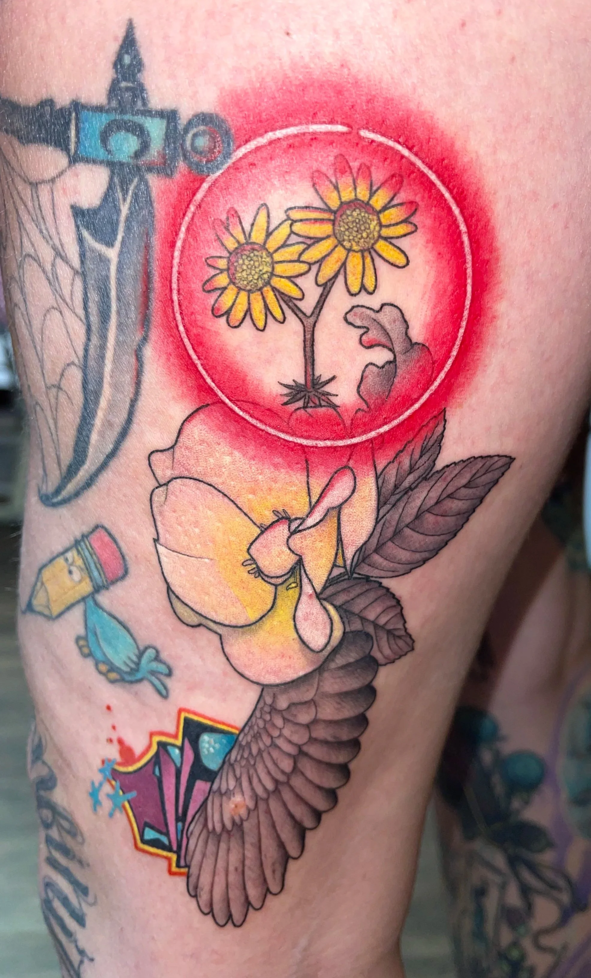 Tattoo of a starting circle with two yellow daisies inside, a blooming flower with yellow petals, leaves, and an eagle wing beneath, on a person's arm.