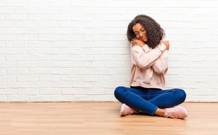 Smiling woman sitting on wooden floor with crossed legs, hugging herself against a white brick wall.