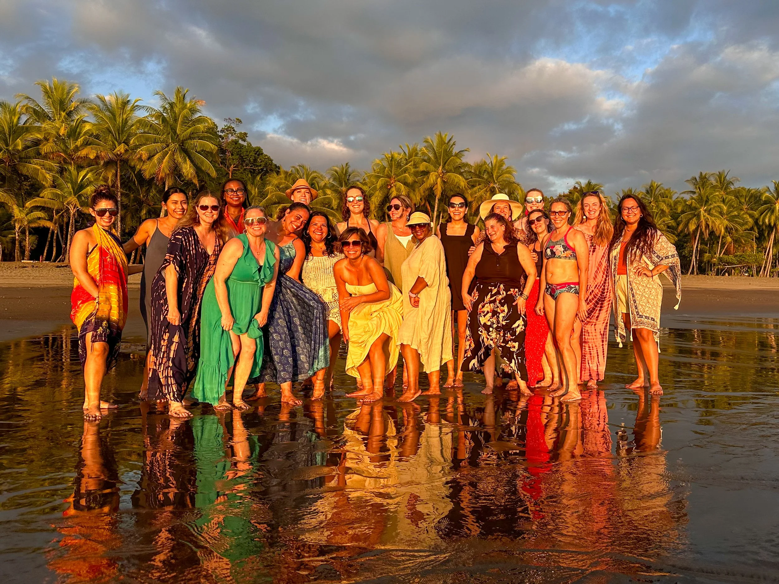 A group of women standing on a beach with palm trees in the background, smiling and enjoying the sunset.
