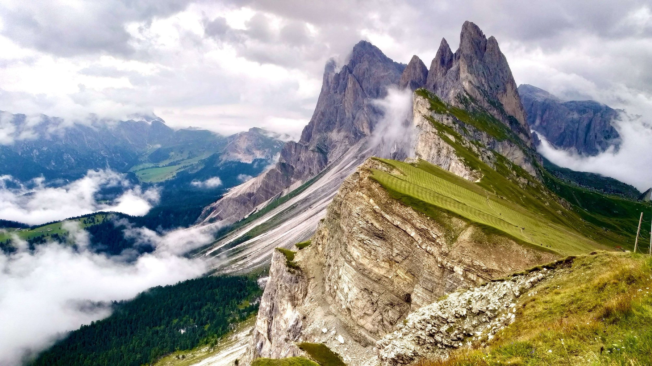 Mountain landscape with steep rocky peaks, green fields, and clouds surrounding the mountains.
