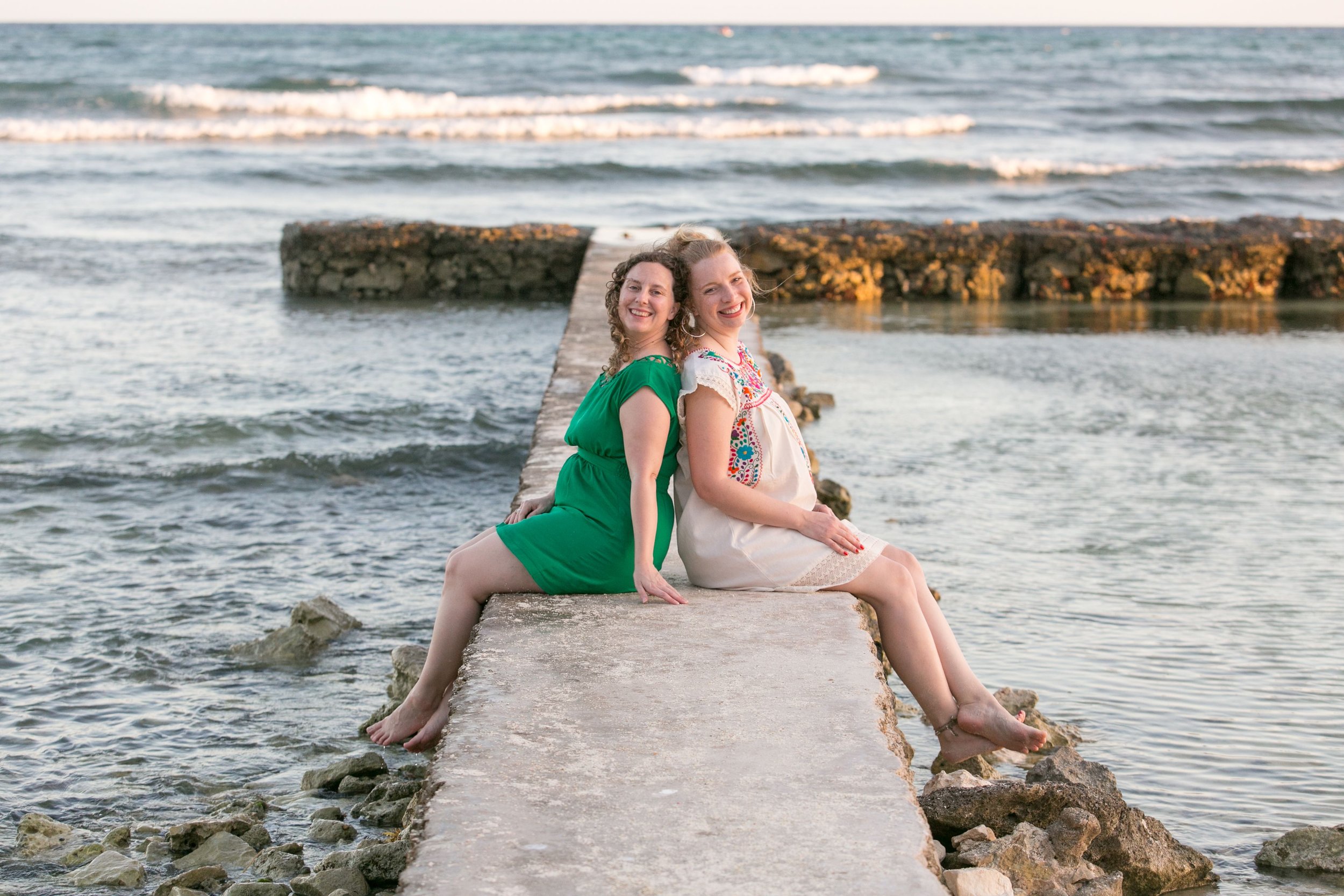 Two women sitting on a concrete pier by the water, smiling, with ocean waves in the background during sunset or sunrise.