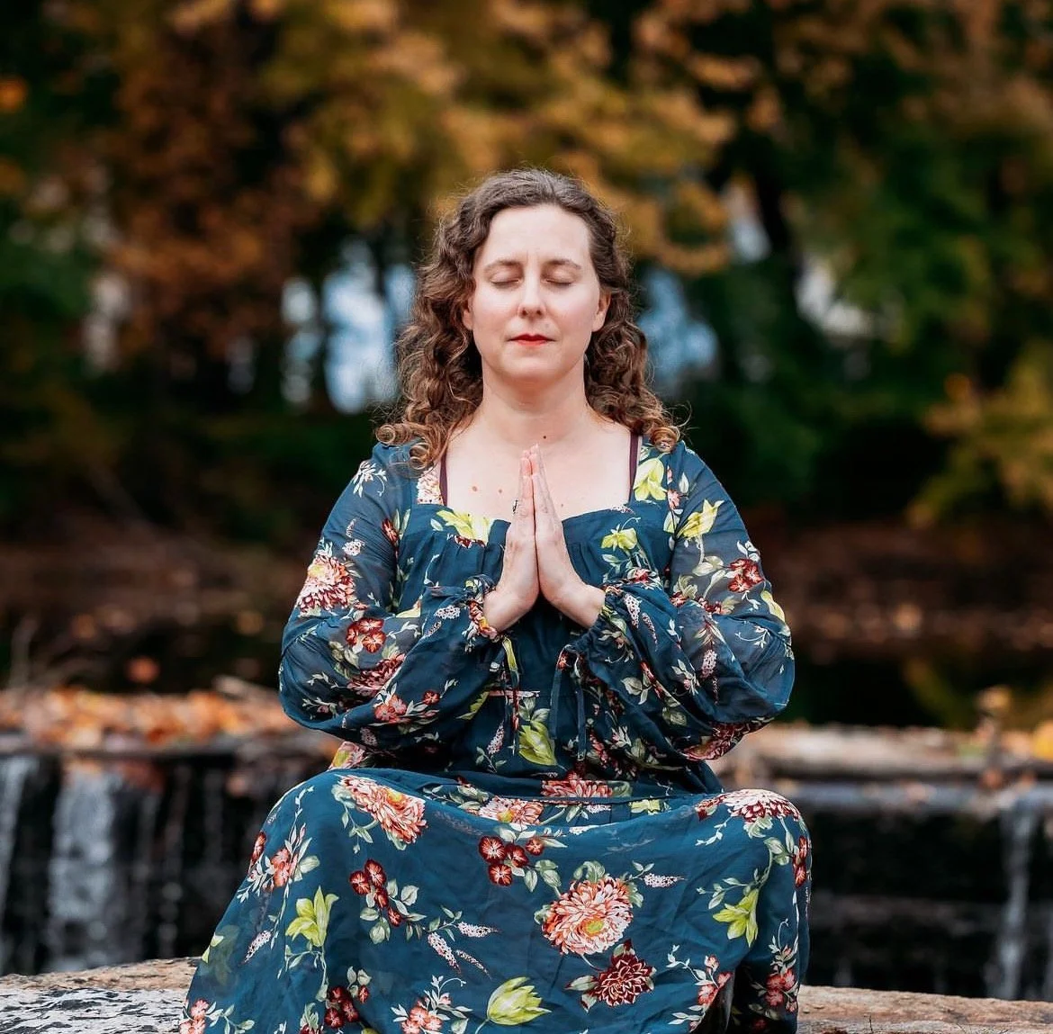 A woman sitting cross-legged outdoors in a meditative pose with hands pressed together in front of her chest, eyes closed, surrounded by autumn trees.