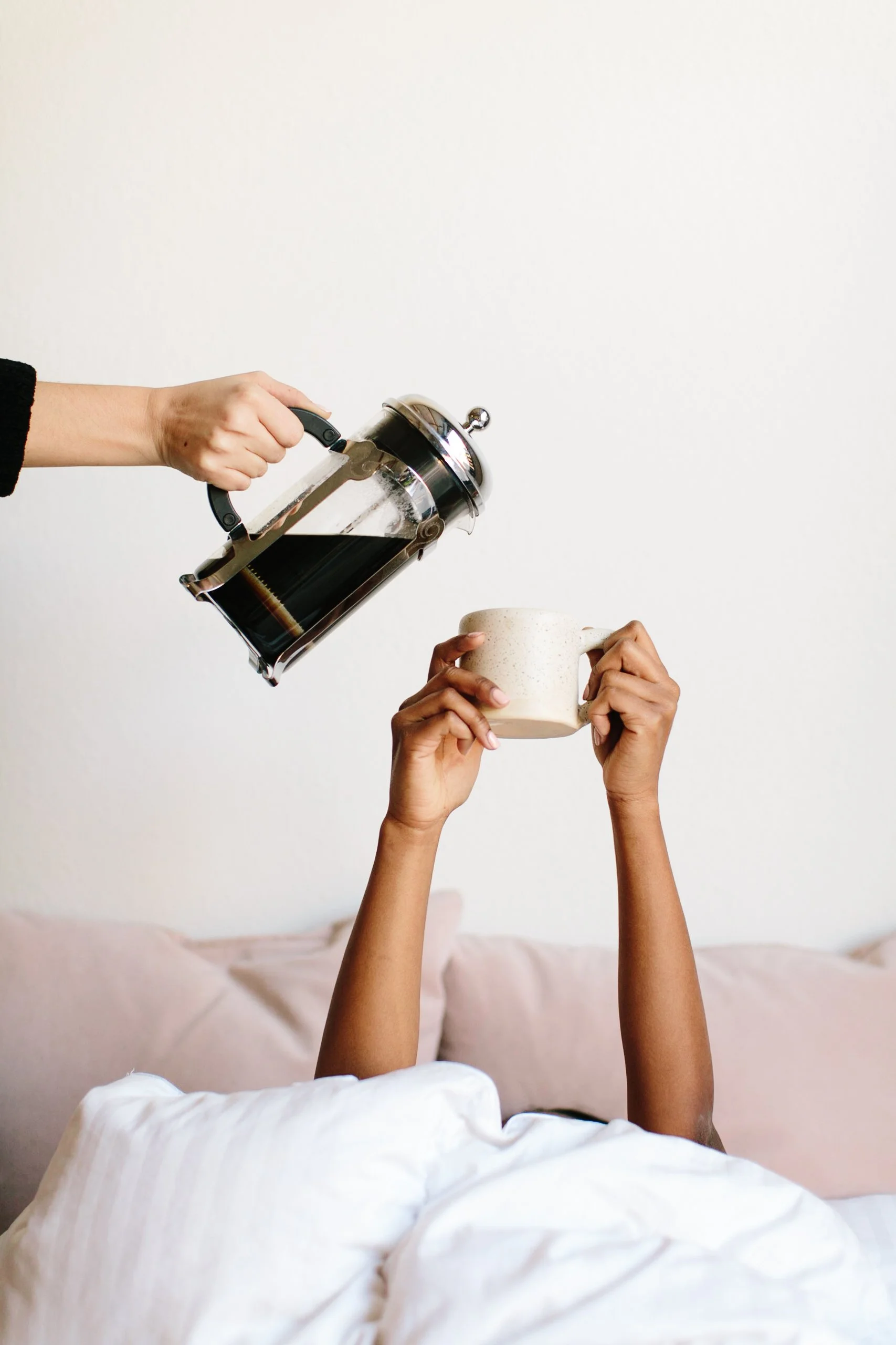 Black woman in bed mostly hidden under covers, reaching a mug up overhead. Another woman, white, stands outside the photo frame so only her forearm and hand shows, pouring coffee into the mug.