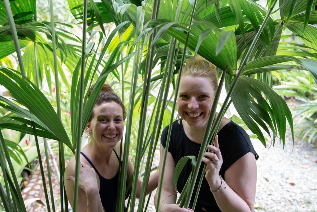 Two women smiling and posing among green tropical plants outdoors.