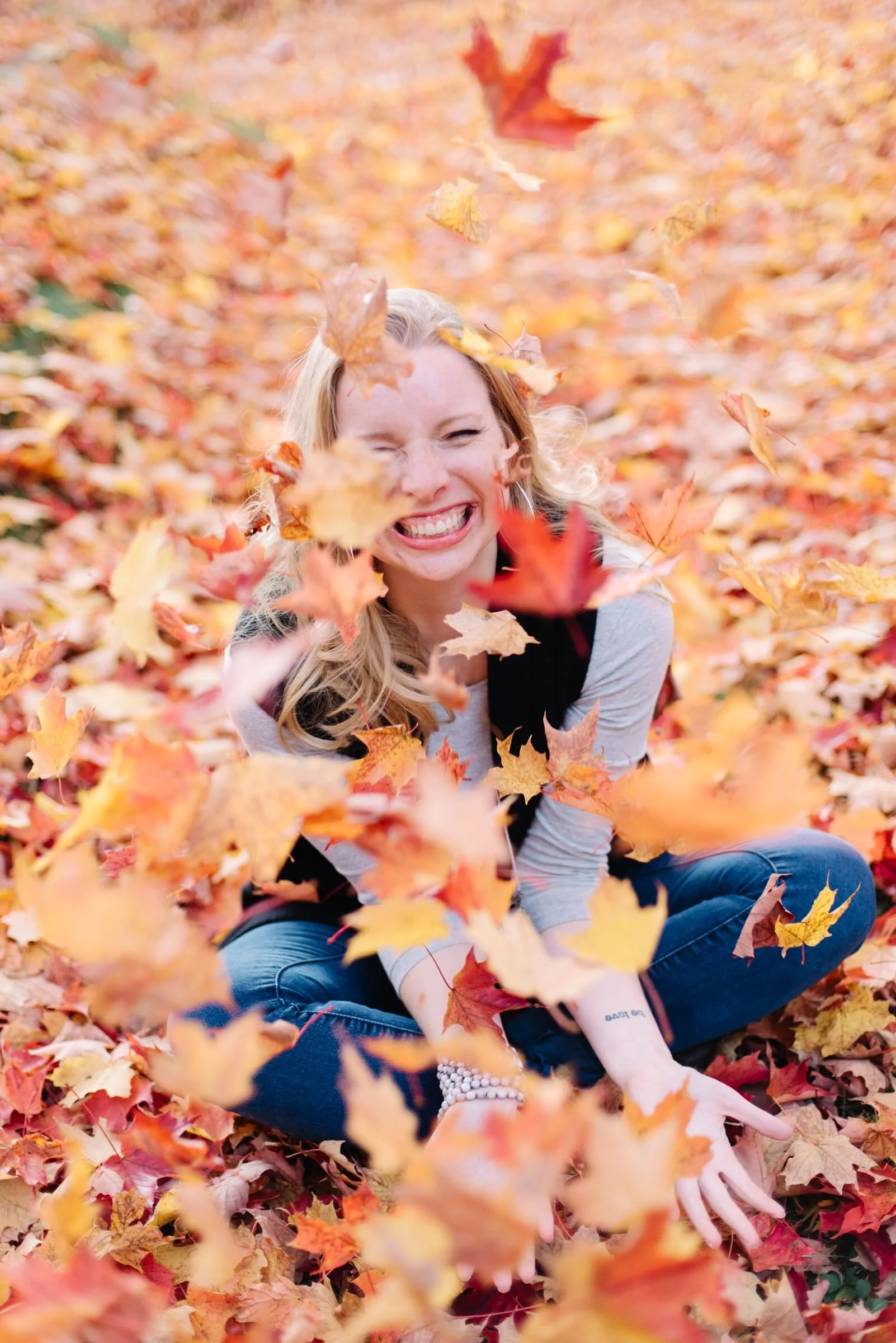 A woman sitting in a pile of orange and yellow fall leaves, smiling and throwing leaves in the air during autumn.