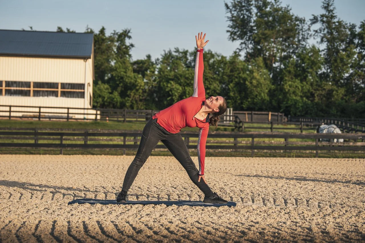 Woman performing a side stretch exercise on a yoga mat outdoors on a sunny day. She is wearing a red long-sleeve top and black pants, with trees and a building in the background.
