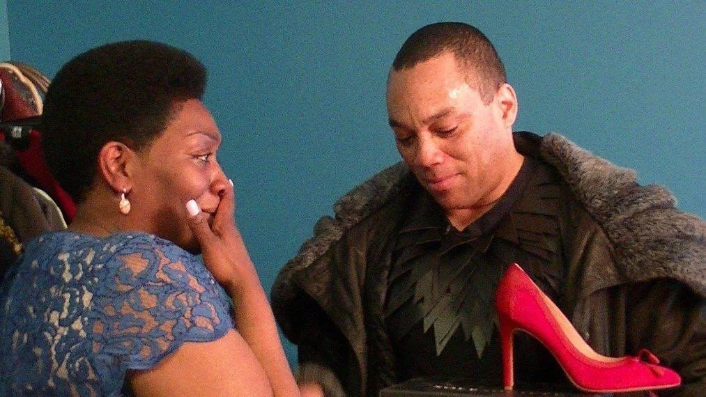 Against a teal wall, a man and a tearful woman look down at a red heel. The woman has a visible tear on her cheek as she gazes at the shoe.