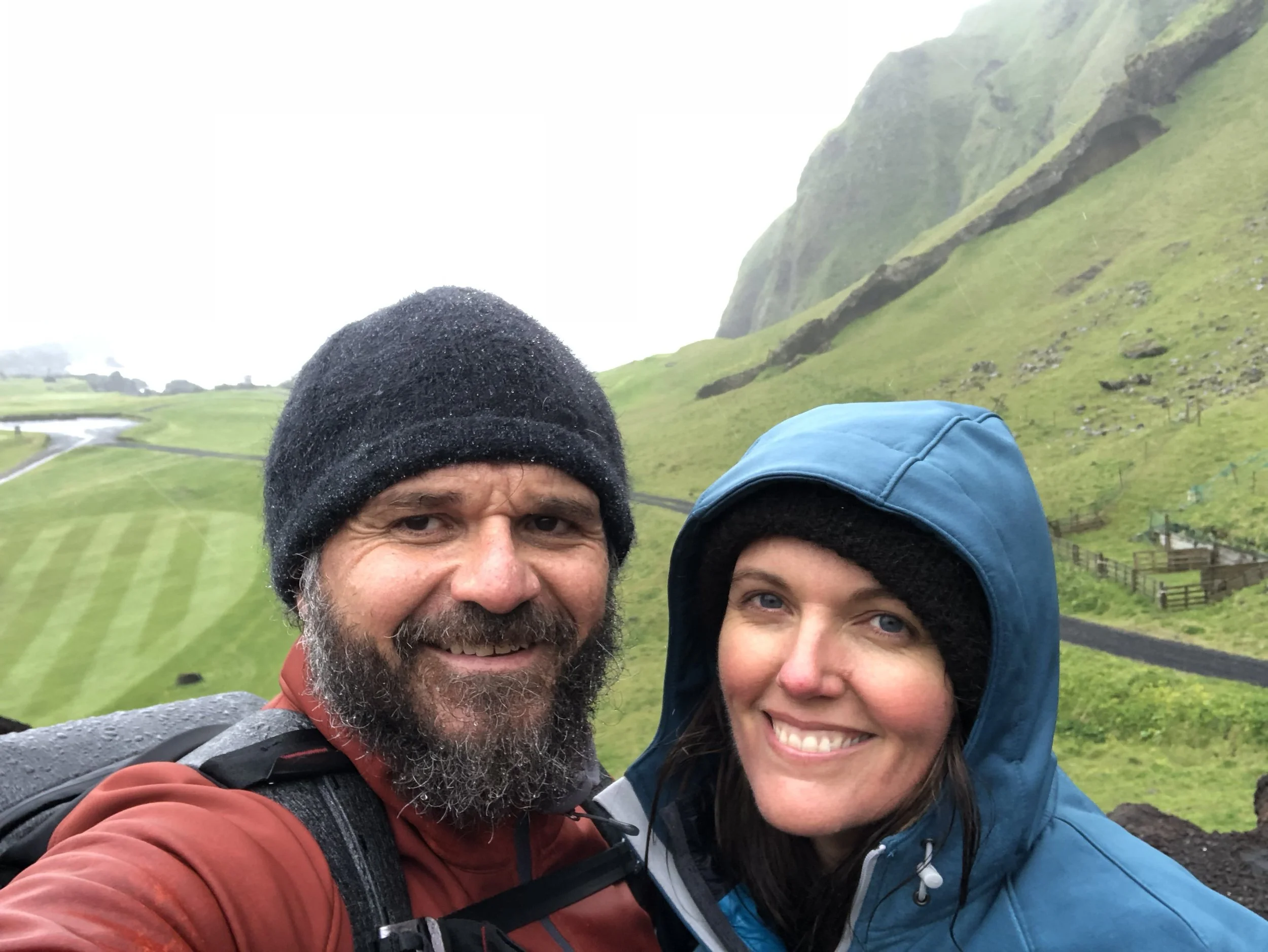 Smiling couple in winter hiking gear taking selfie with green hills in background.