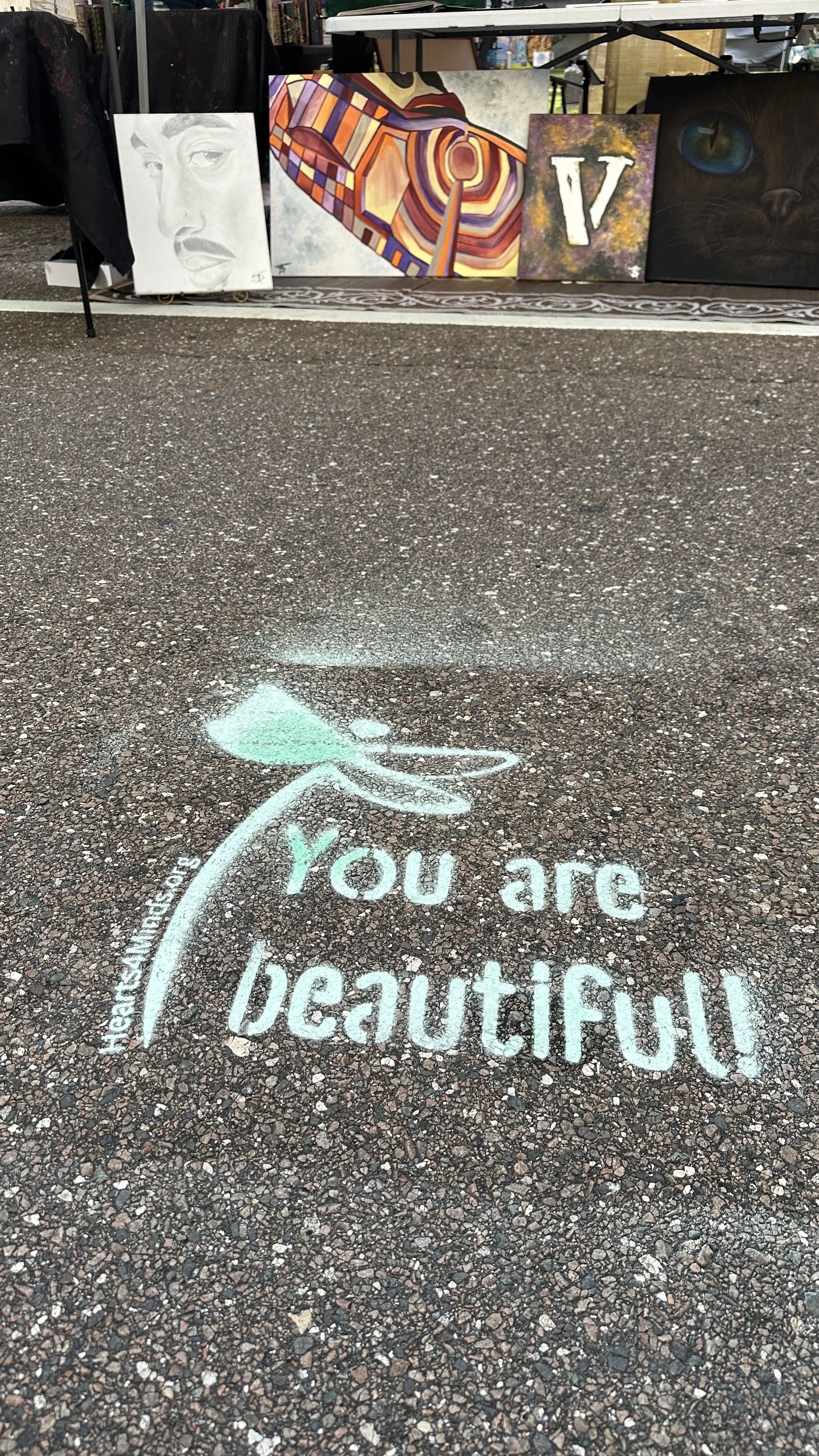 Turquoise chalk art on pavement reading 'You are beautiful' with dragonfly symbol and artist canvases in background.