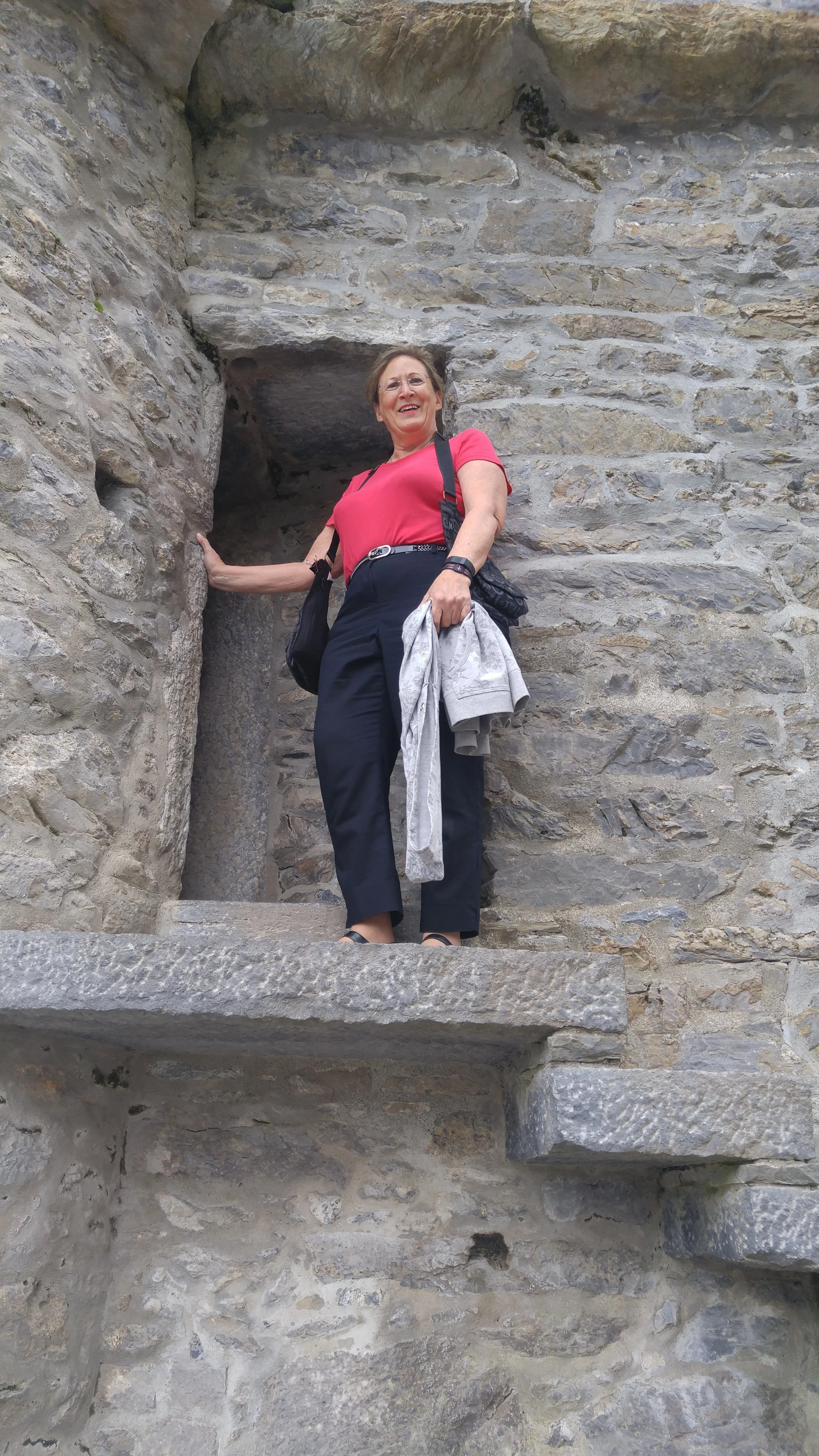 Smiling woman in red top and black pants standing on stone steps in historic stone structure.
