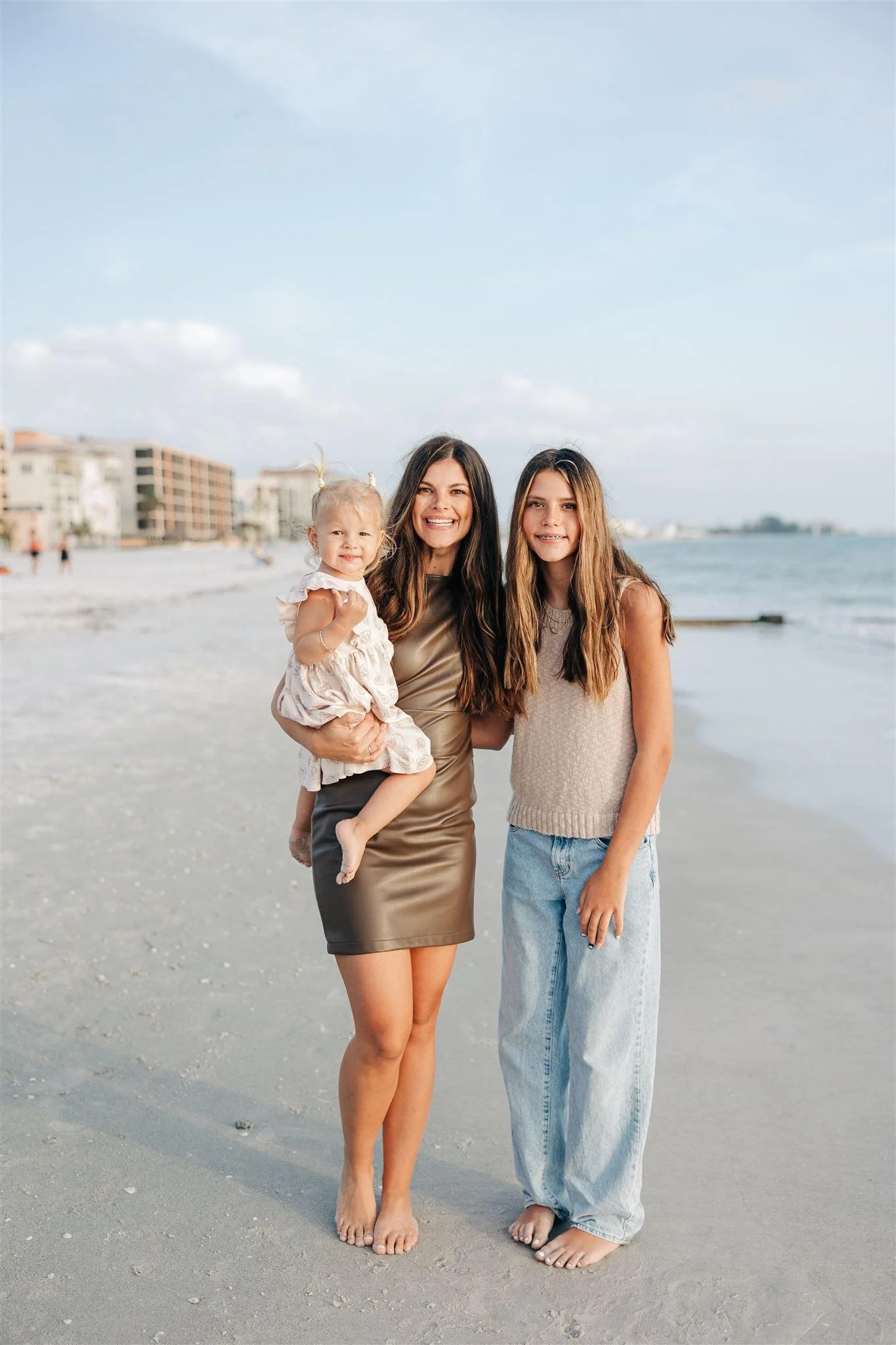 A mother and 2 children standing on the beach smiling with the ocean in the background.