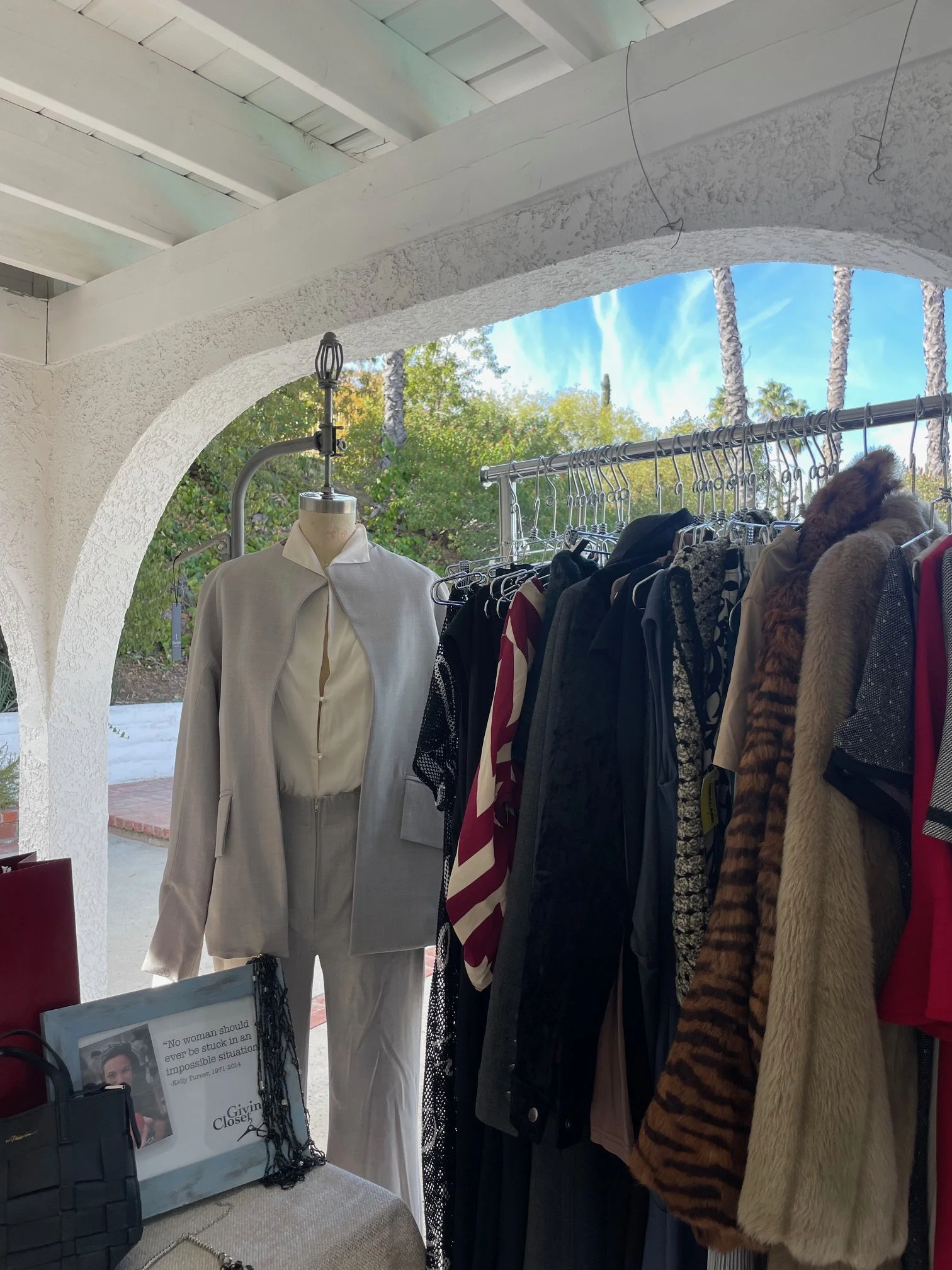 A clothing display under a white archway featuring a mannequin in a grey suit and a rack of various garments, including a faux-fur coat.