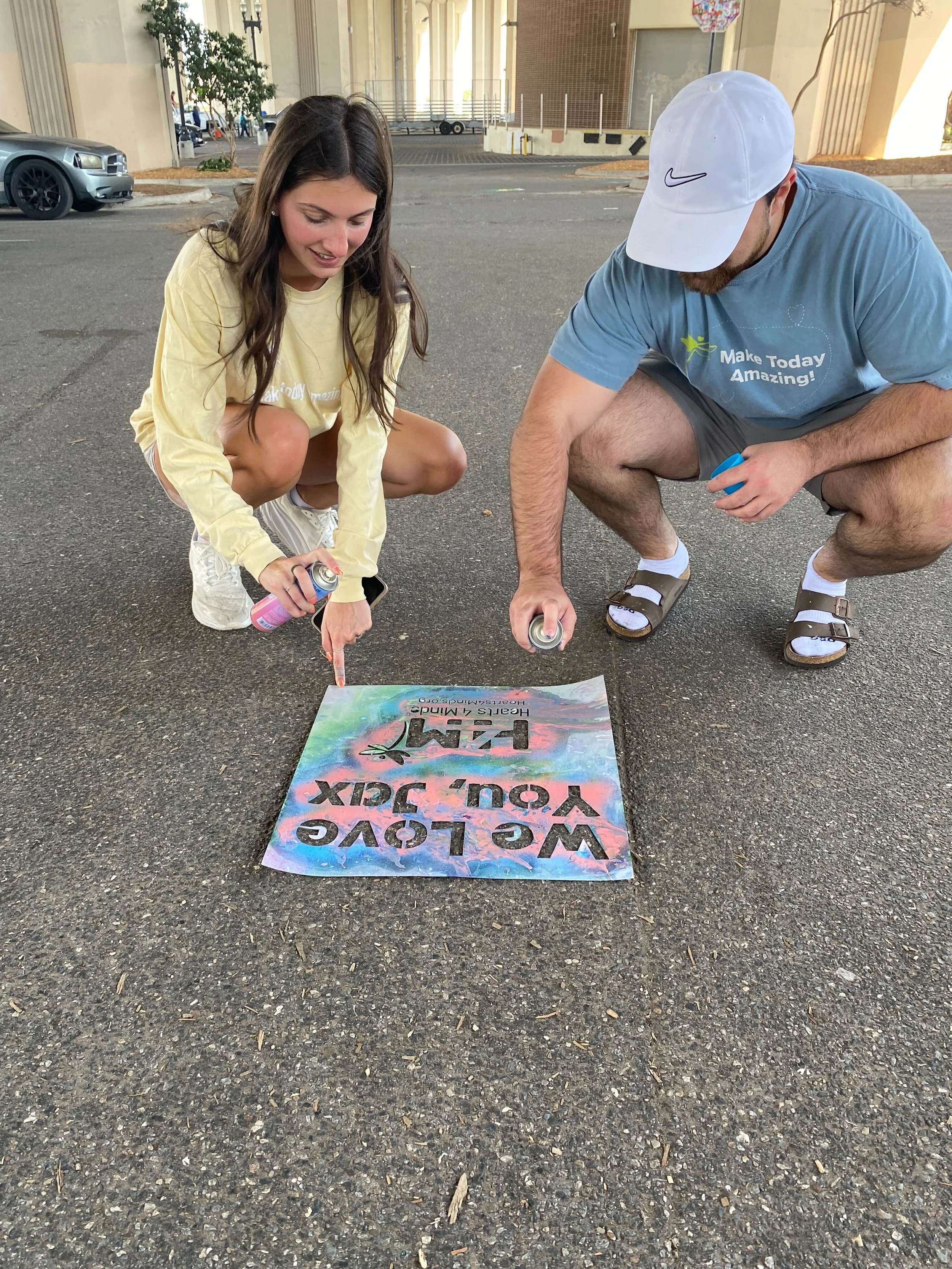 Woman in yellow shirt and man in blue 'Make Today Amazing' shirt kneeling on pavement spray painting a vibrant tie-dye style Hearts 4 Minds sign that reads 'We Love You, Jax'.