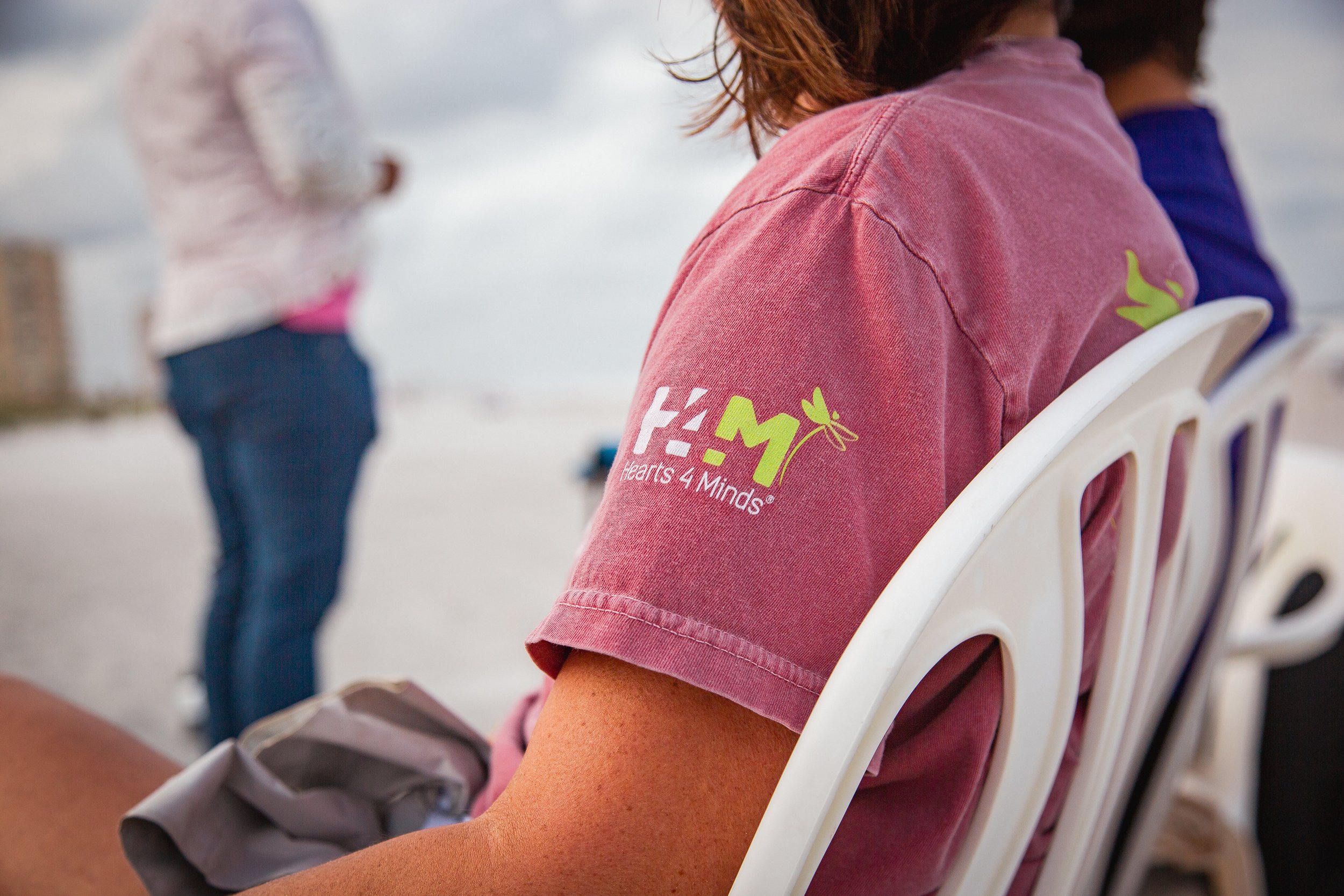 Person wearing pink Hearts 4 Minds branded t-shirt sitting in white chair at outdoor community event