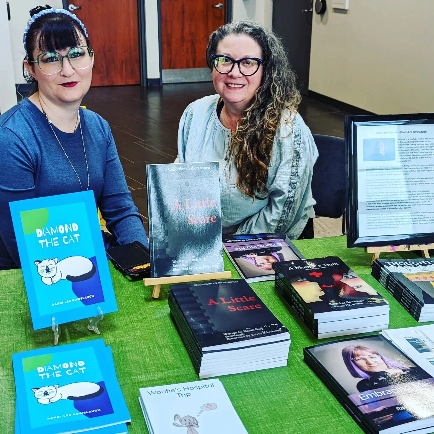 Two women sit in front of a table with a dozen books piled on smiling at the camera.