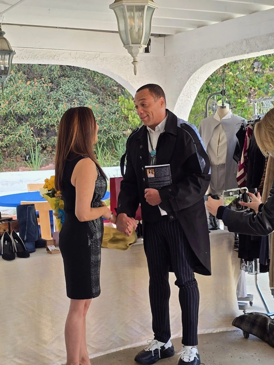A man in a black coat holding a book and holding hands with a woman in a black dress at an outdoor event.
