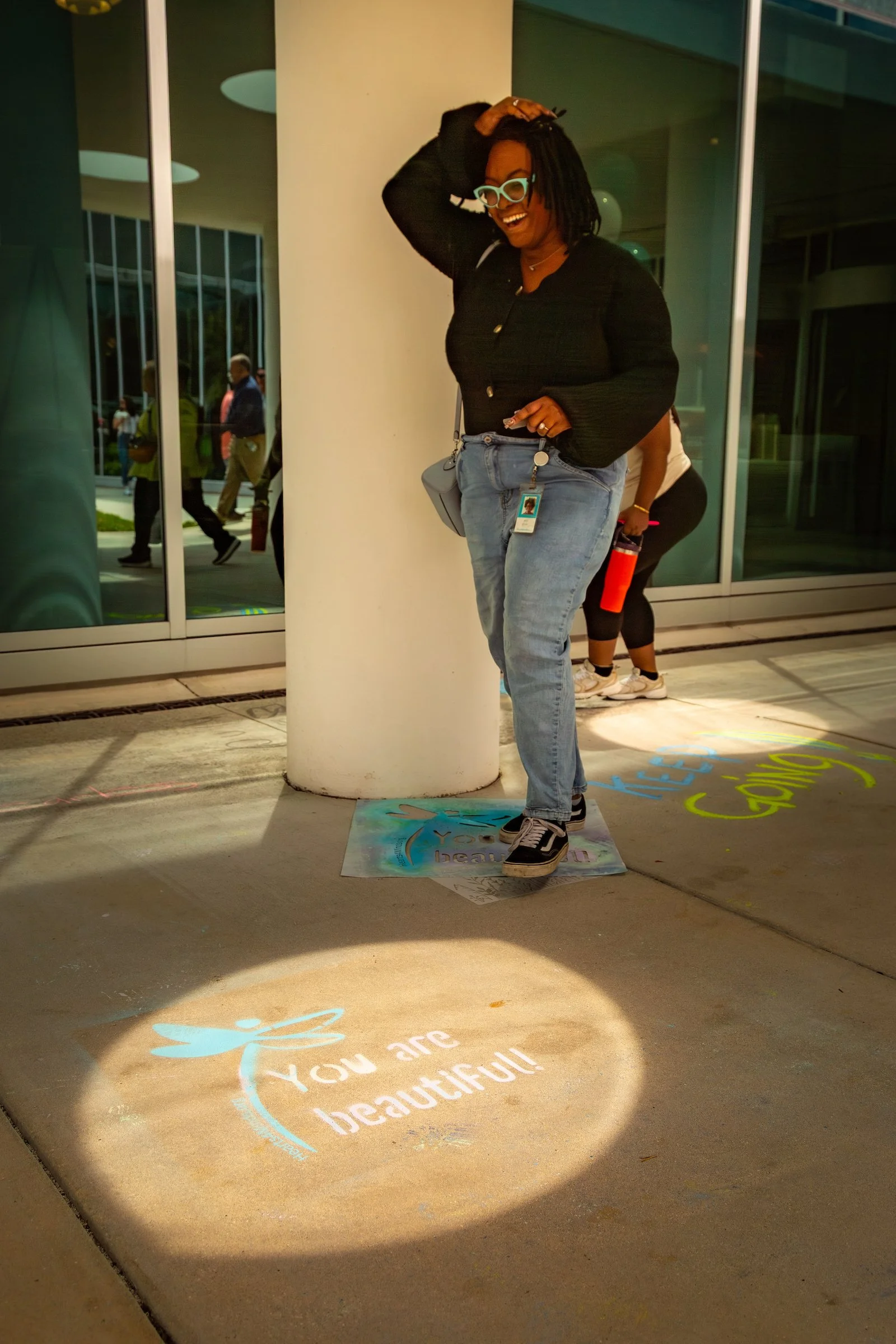 Smiling woman stands beside sidewalk chalk art with a dragonfly and the message "You are beautiful!" at a Hearts 4 Minds event.