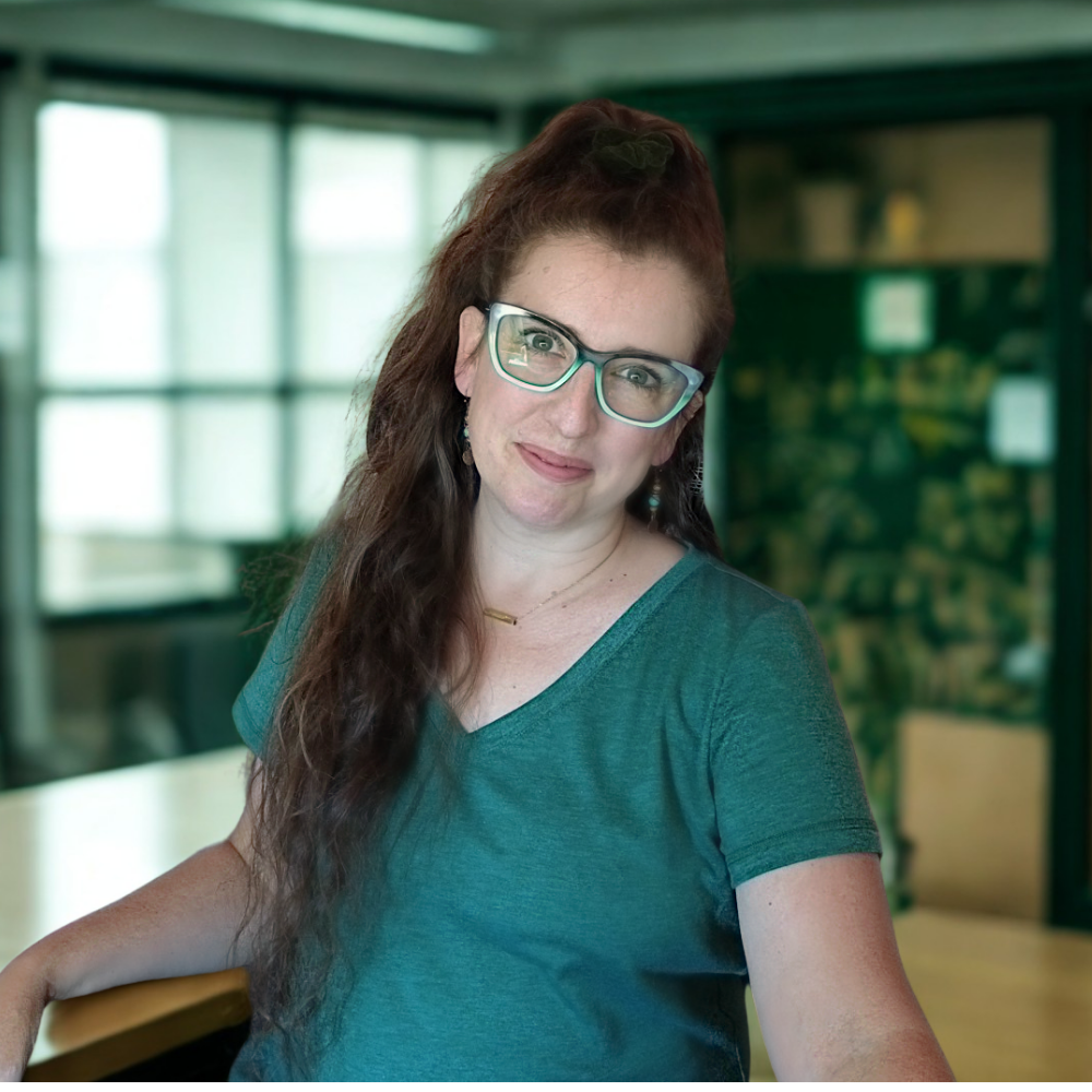 Smiling woman with long brown hair wearing teal-framed glasses and a teal v-neck shirt in a bright indoor setting with windows.