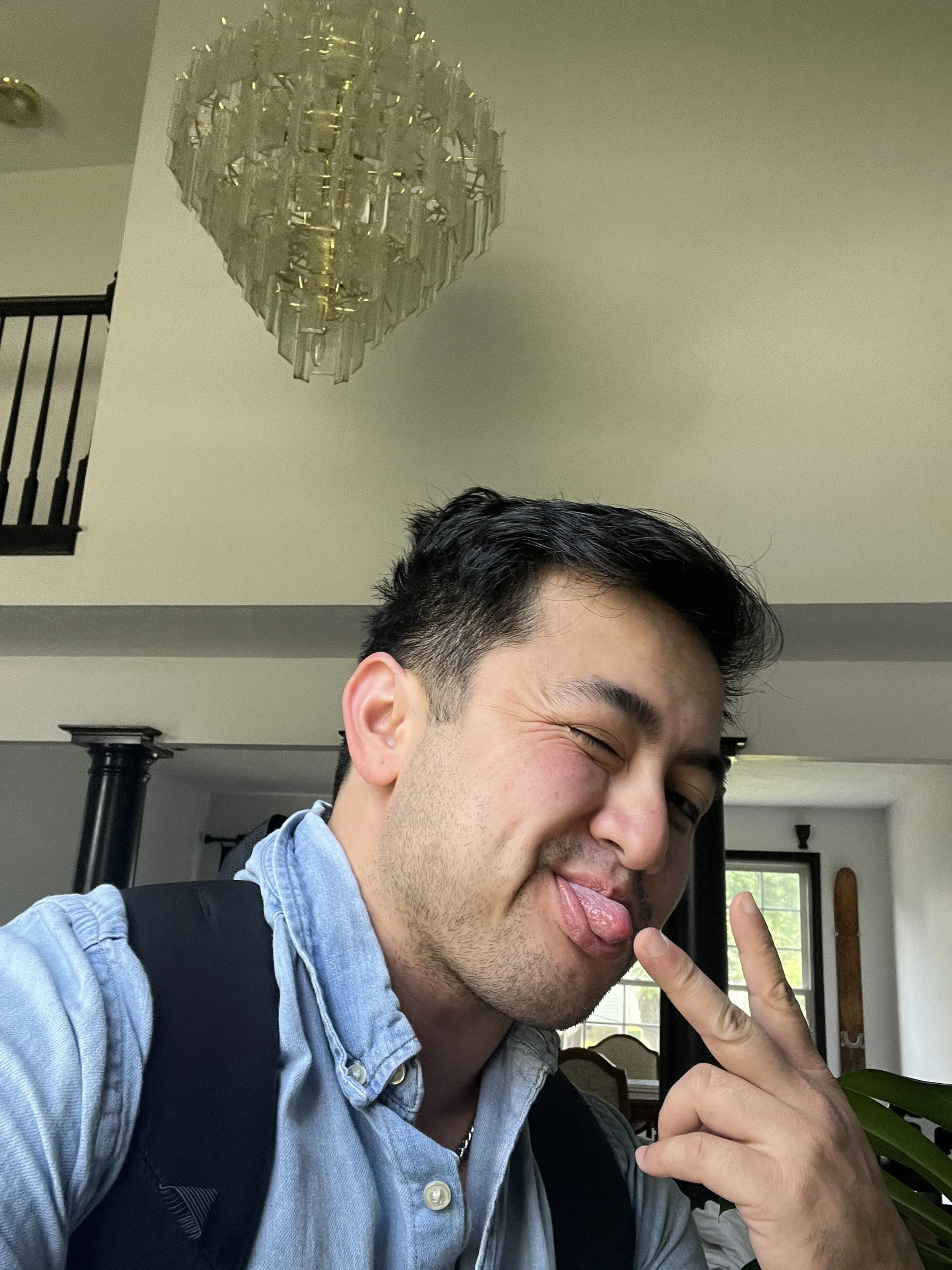 Young man taking a selfie in a well-lit room, making a playful face with tongue out and holding up two fingers in a peace sign.