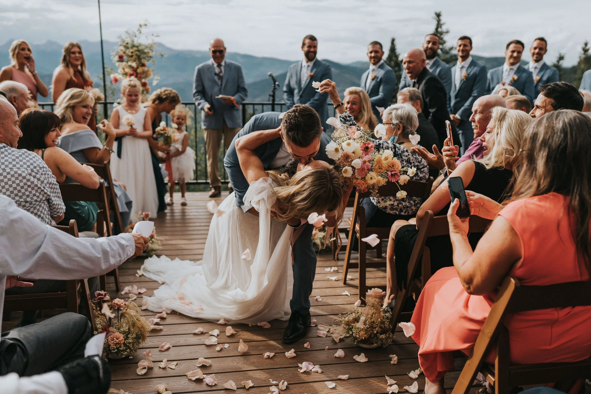 A couple kissing during a wedding ceremony on a wooden outdoor deck, surrounded by friends and family with mountains in the background.