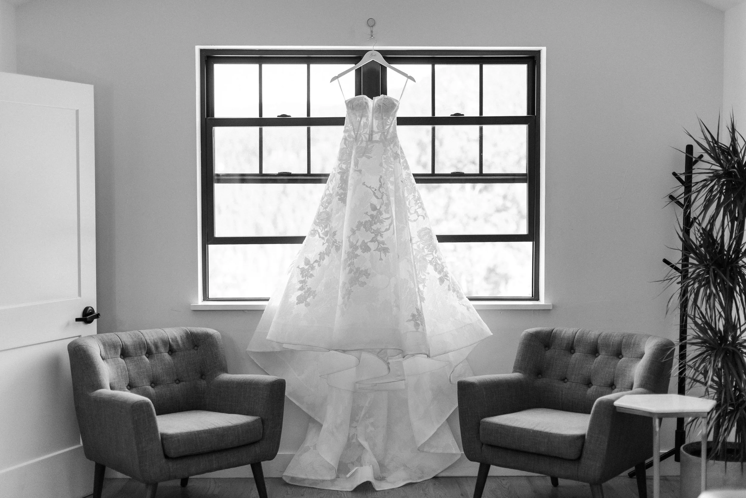 A black and white photo of a beautiful princess style wedding dress over a window with a wide, flowing train, and leaves and flowers across the body.