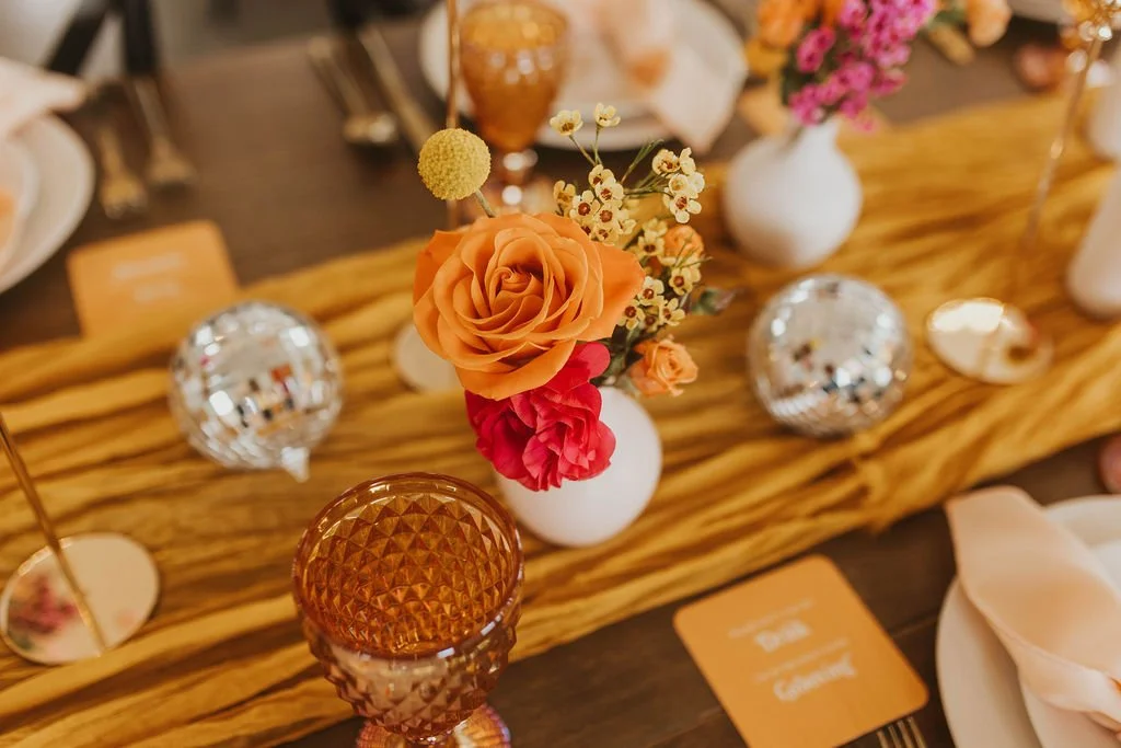 A table centerpiece with a small bouquet of orange, pink, and white flowers in a white vase on a wooden table runner, surrounded by decorative glass ornaments, candles, and place cards.