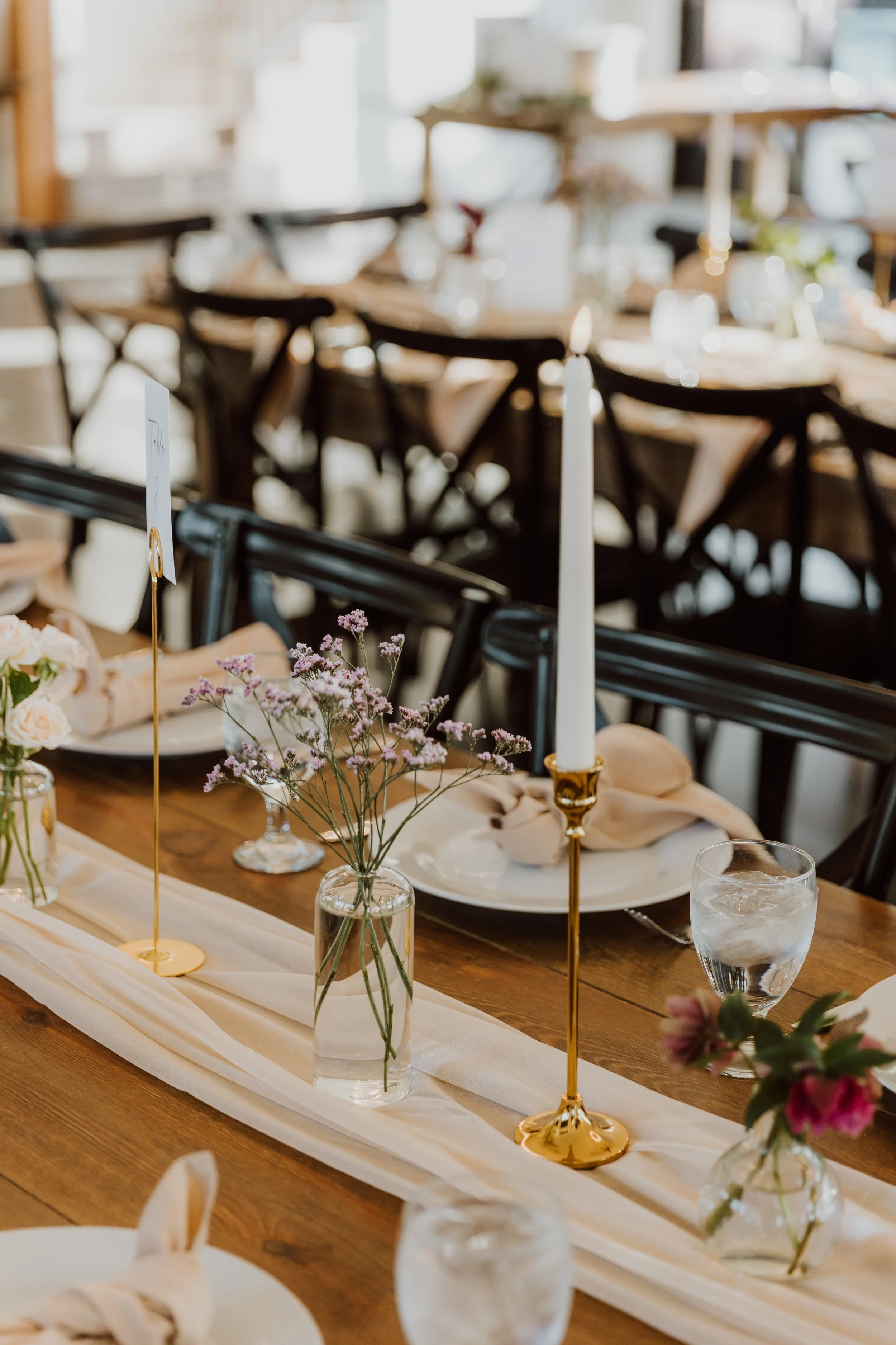 A decorated dinner table with vases of flowers and tall white candles, set for a celebration or event in a well-lit room.