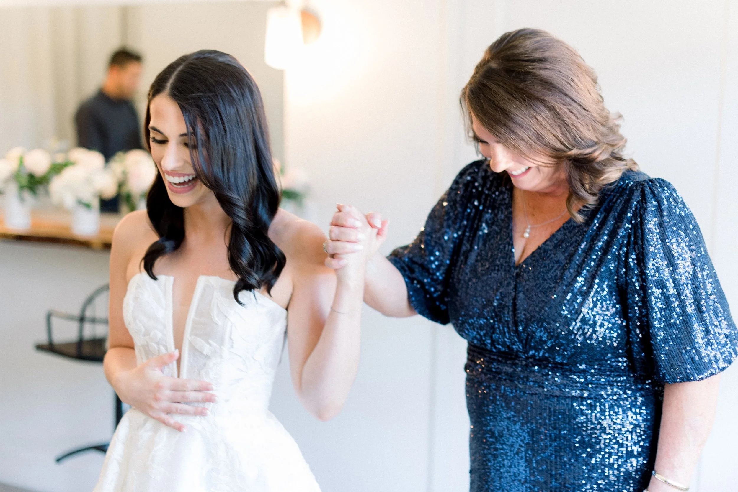 A mother and daughter hold hands and smile while getting ready on her wedding day.