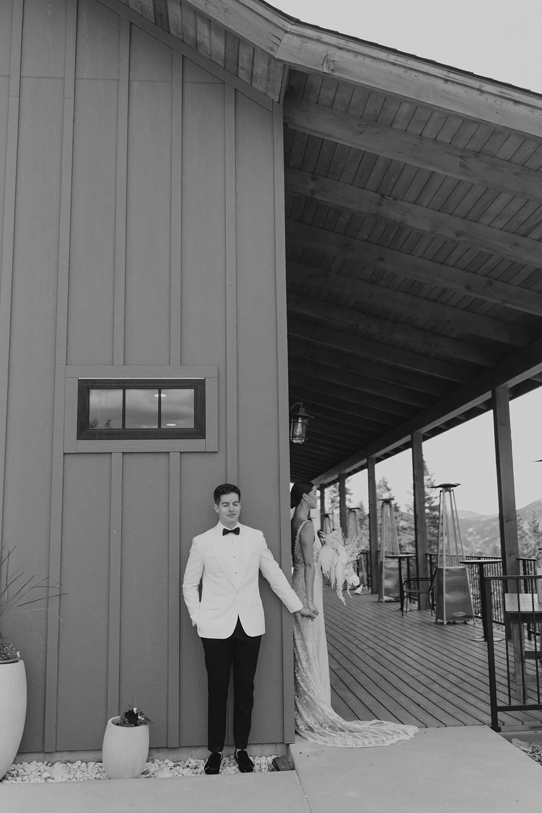A black and white photo of a couple in wedding attire, standing outside a building with a wooden exterior and balcony. The groom, in a tuxedo, stands with his eyes closed and holding the bride's hand, who is wearing a lace wedding gown and holding a bouquet, looking away.