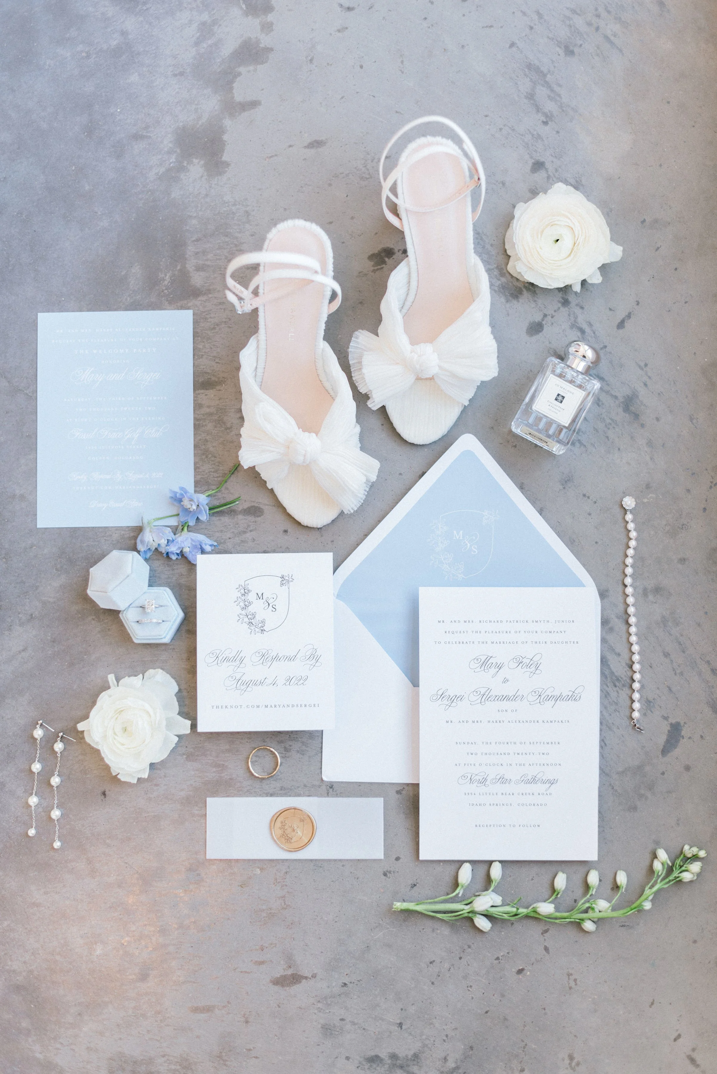 A flat lay image of wedding details with ruffly white heels, perfume, and white and blue wedding invitations for a sleek, clean, timeless mountain wedding.