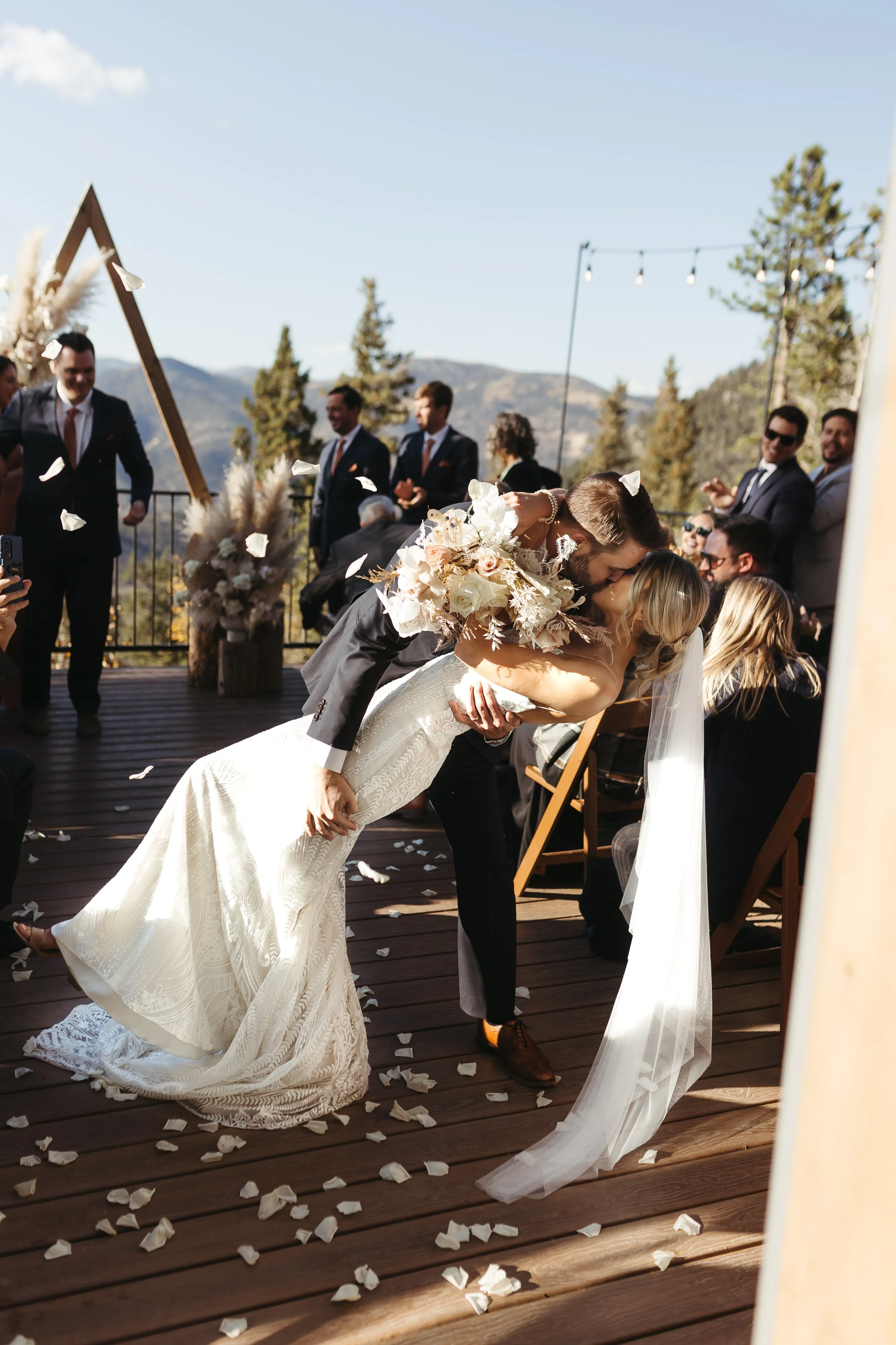 A deep dip kiss at the end of the aisle on a wedding day with beautiful white flower petals tossed in the air.