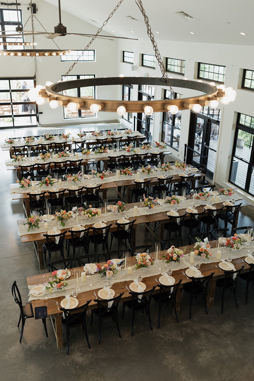 Empty banquet hall with long wooden tables decorated with pastel-colored flowers, candles, and white plates, illuminated by a circular chandelier with exposed bulbs.