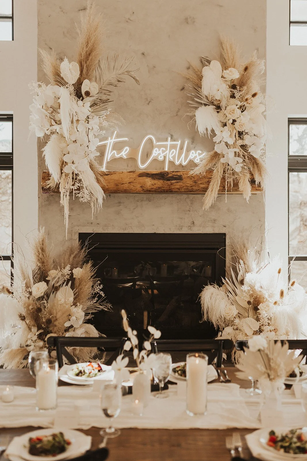 Decorated fireplace with a wooden mantel, floral arrangements with white flowers and pampas grass, and a neon sign reading 'The Costellos' in cursive font.